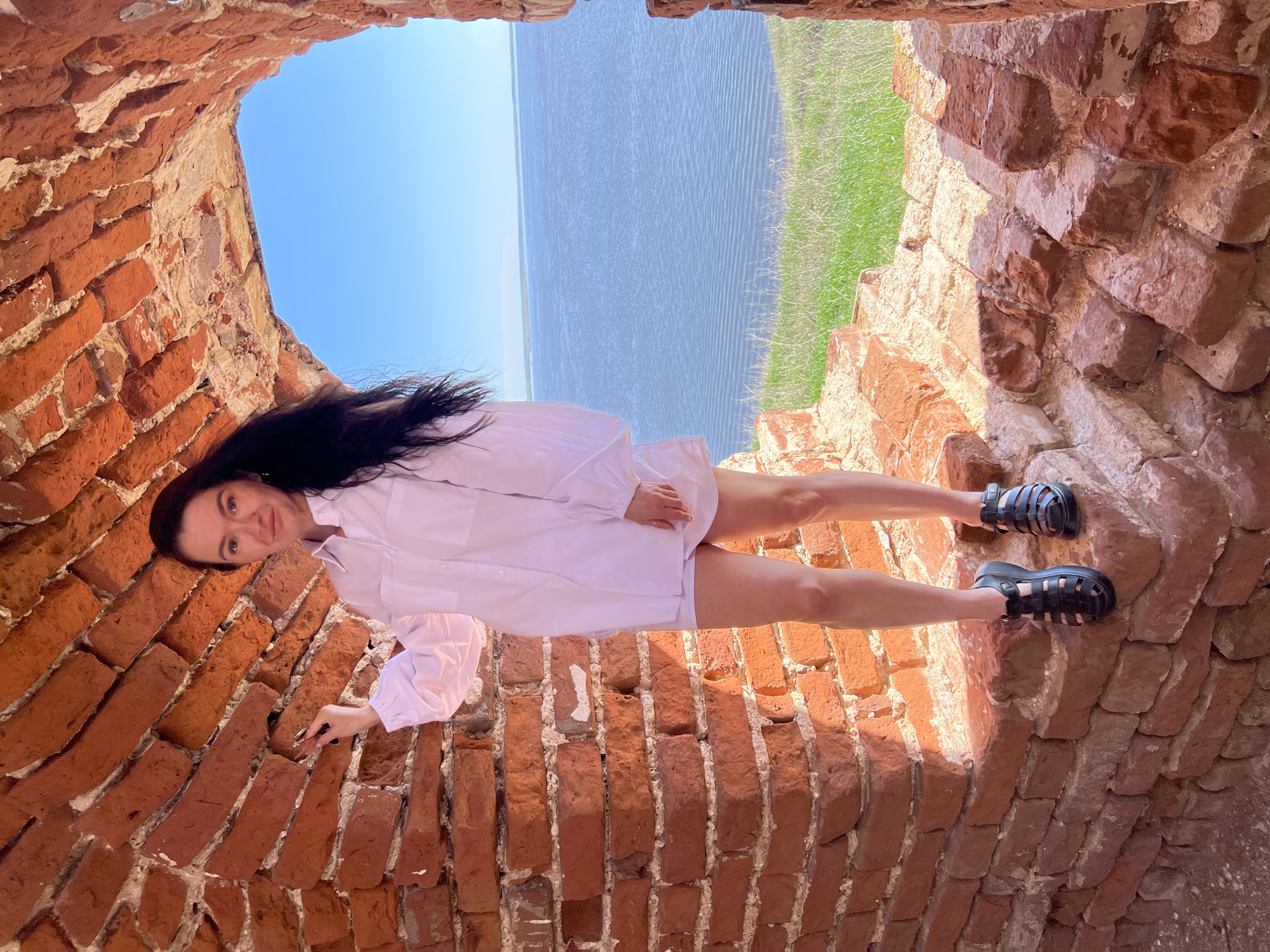 A woman stands inside a brick structure in Шуран, Russia. The structure has a weathered red brick interior and looks out onto a body of water with a grassy bank on the opposite side. The woman has long dark hair and is wearing a white shirt-dress and black sandals. The sky above is blue.