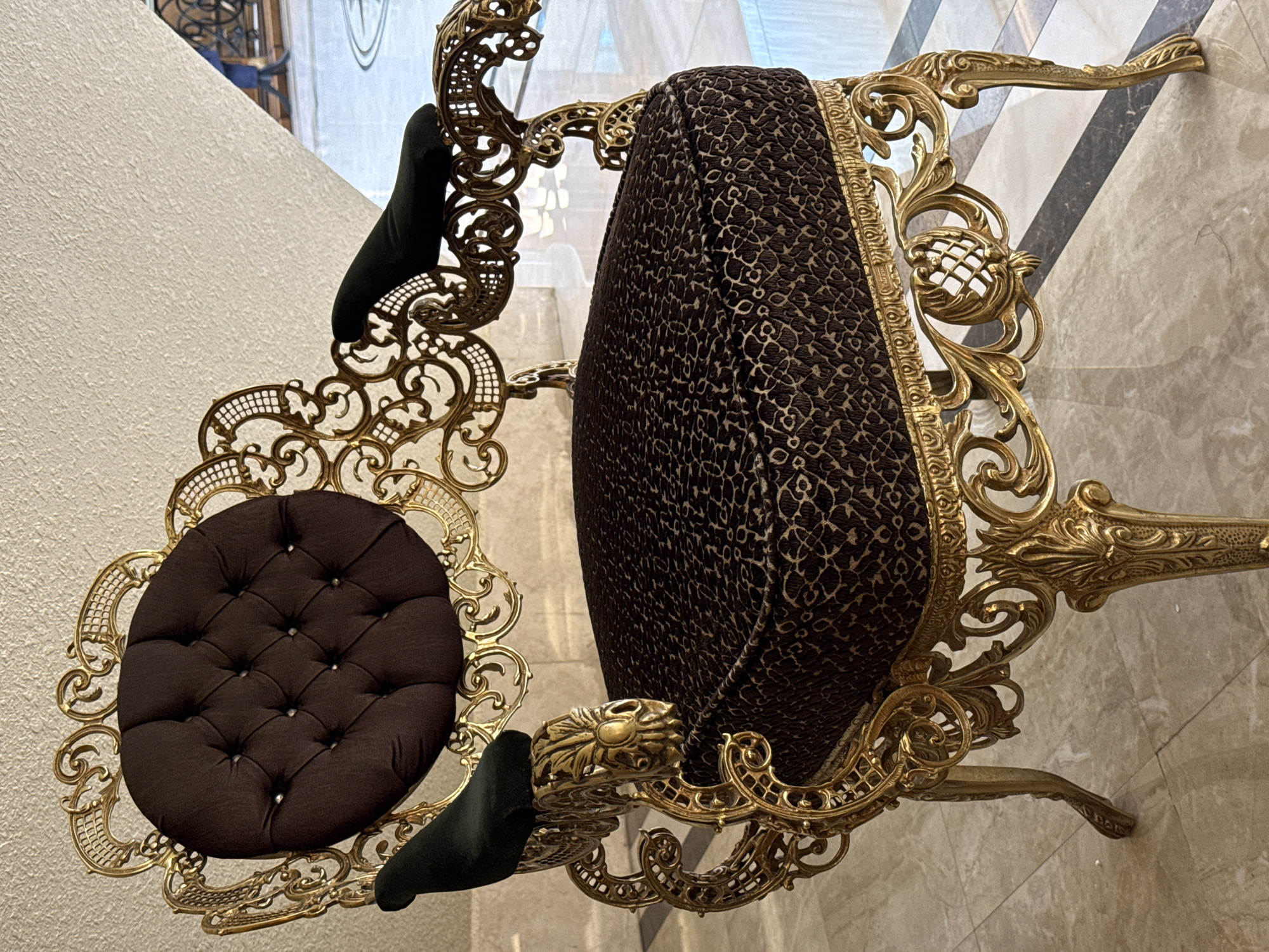 A close-up, eye-level shot shows an ornate, gold-gilt armchair with dark brown tufted upholstery on the backrest and a patterned dark brown cushion on the seat. Dark green velvet armrests are affixed to the elaborate gold frame. The chair is positioned on a tiled floor with a geometric pattern of beige, white, and grey tiles. In the background, through a glass partition, a metal railing and the blurry figures of two individuals can be seen. The overall impression is one of luxury and opulence, likely within a grand interior space. This image was taken in Baku, Azerbaijan.