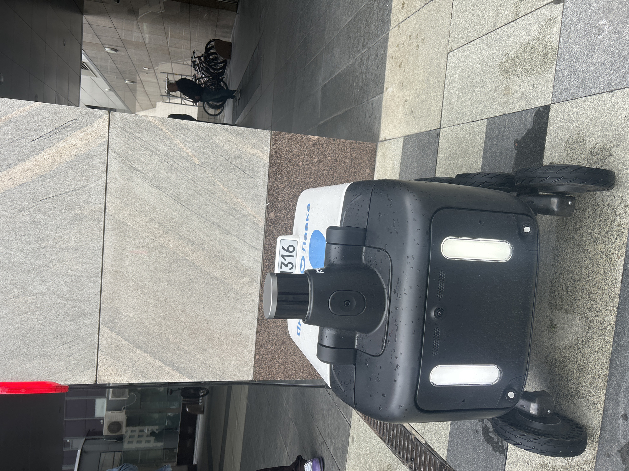 A high-angle medium shot shows an autonomous delivery robot on a stone-tiled sidewalk near a building. The robot is predominantly black and white, with a cylindrical camera unit mounted on top. A license plate with the number "316" is visible on the robot's front. The robot has four small wheels.
The environment includes building walls constructed of large stone blocks, a bicycle rack with multiple bicycles in the background, and a storm drain along the edge of the sidewalk. Reflections in the building windows suggest an urban environment with buildings and other structures across the street. The location is identified as Unknown, Unknown.