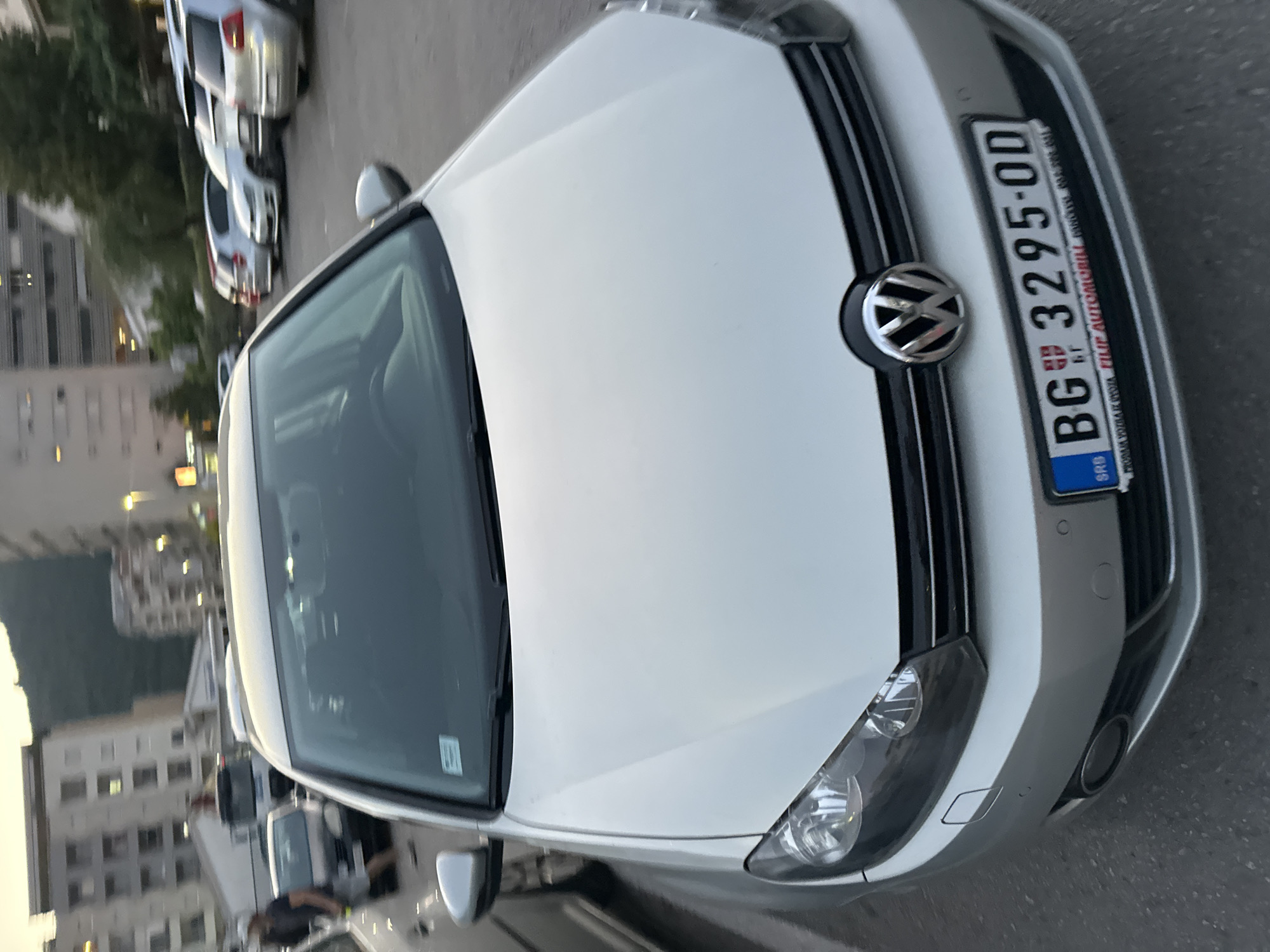 A silver Volkswagen Golf is parked in an outdoor parking lot in Budva, Montenegro. The car is facing the viewer, with its front grille, headlights, and license plate clearly visible. The license plate reads "BG 3295-OD". In the background, there are several other parked cars, a multi-story building, and trees. The sky is overcast, suggesting it is either early morning or late afternoon. The scene appears to be a typical urban parking area.