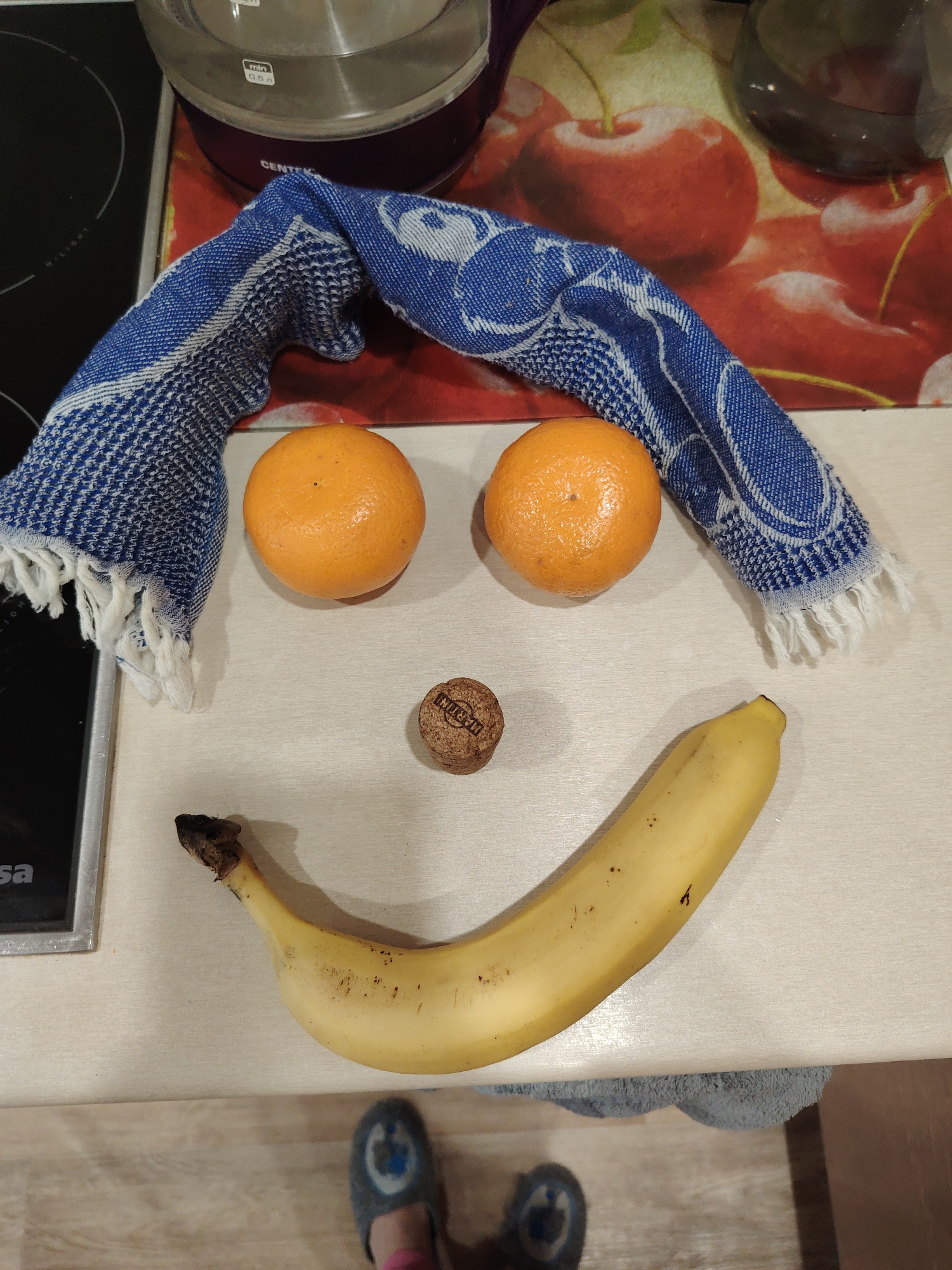 The image depicts a whimsical arrangement of objects on a light-colored countertop, resembling a face. Two oranges represent eyes, a champagne cork serves as a nose, and a banana forms a smiling mouth. A blue and white dish towel arches above the oranges, suggesting eyebrows or hair. The countertop also holds a glass kettle on a base, positioned behind the "face," with a cutting board featuring a cherry print below the kettle. Part of a stovetop is visible on the left edge of the image. Below the countertop, the lower portion of a person's feet wearing slippers can be seen. The location is Unknown, Unknown.