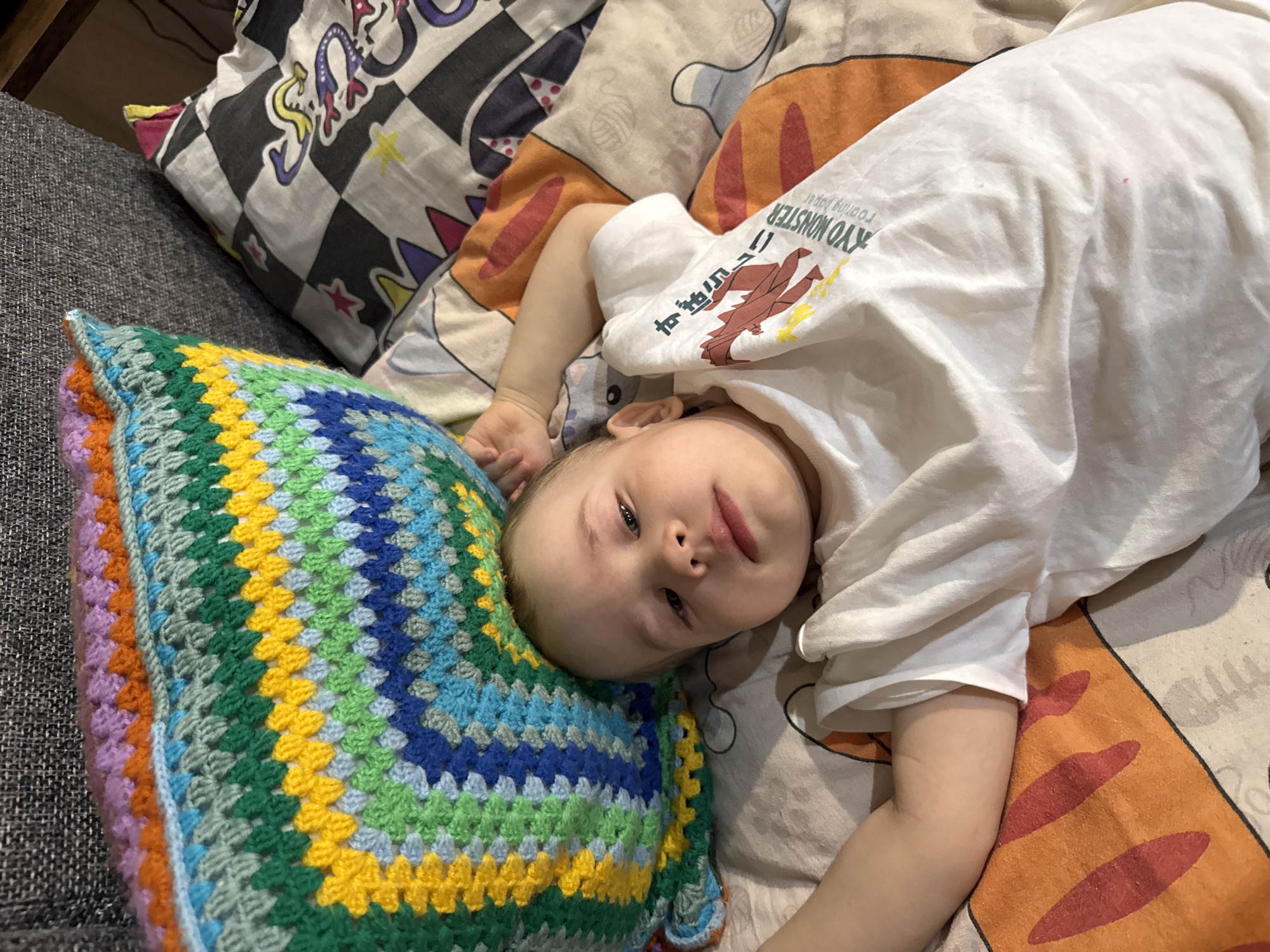 A baby lies on a bed. The baby wears a white shirt with Japanese writing and a cartoon graphic. The baby's head rests on a colorful crocheted pillow. Beside the baby is a black and white checkered pillow. The bed has a patterned orange and white sheet. The baby looks at the camera. The location is not specified, and the city is unknown.