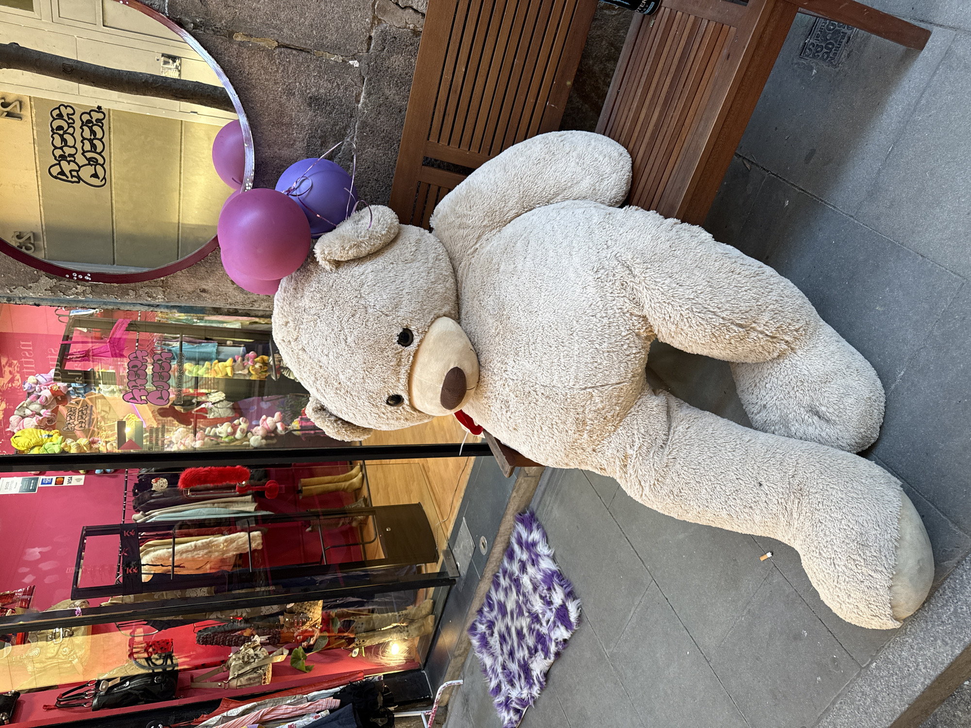 A large teddy bear with a red ribbon around its neck sits on a wooden bench outside a shop in Barcelona, Spain. Two balloons, one pink and one purple, are attached to the bear. A circular mirror reflects graffiti on a building across the street. The shop window behind the bear displays various items for sale, including stuffed animals and clothing. A small purple and white rug lies on the sidewalk near the bear's paw. The sidewalk is made of gray paving stones.