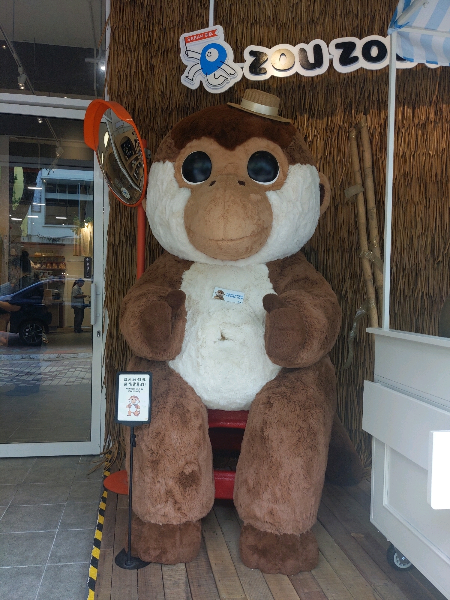 A life-sized stuffed monkey with large eyes, a white chest, and a straw hat is positioned on a wooden platform in front of a wall covered in woven natural material. To the left of the monkey, a reflective orange convex mirror is mounted on a pole. A sign with Chinese and English text stating "Please don't touch me" is placed in front of the monkey. In the background, a glass storefront shows reflections of a city street with a dark car and a person standing near a door with a "PULL" sign. The environment appears to be an outdoor or semi-outdoor setting, possibly a shop entrance in Kota Kinabalu, Malaysia.
