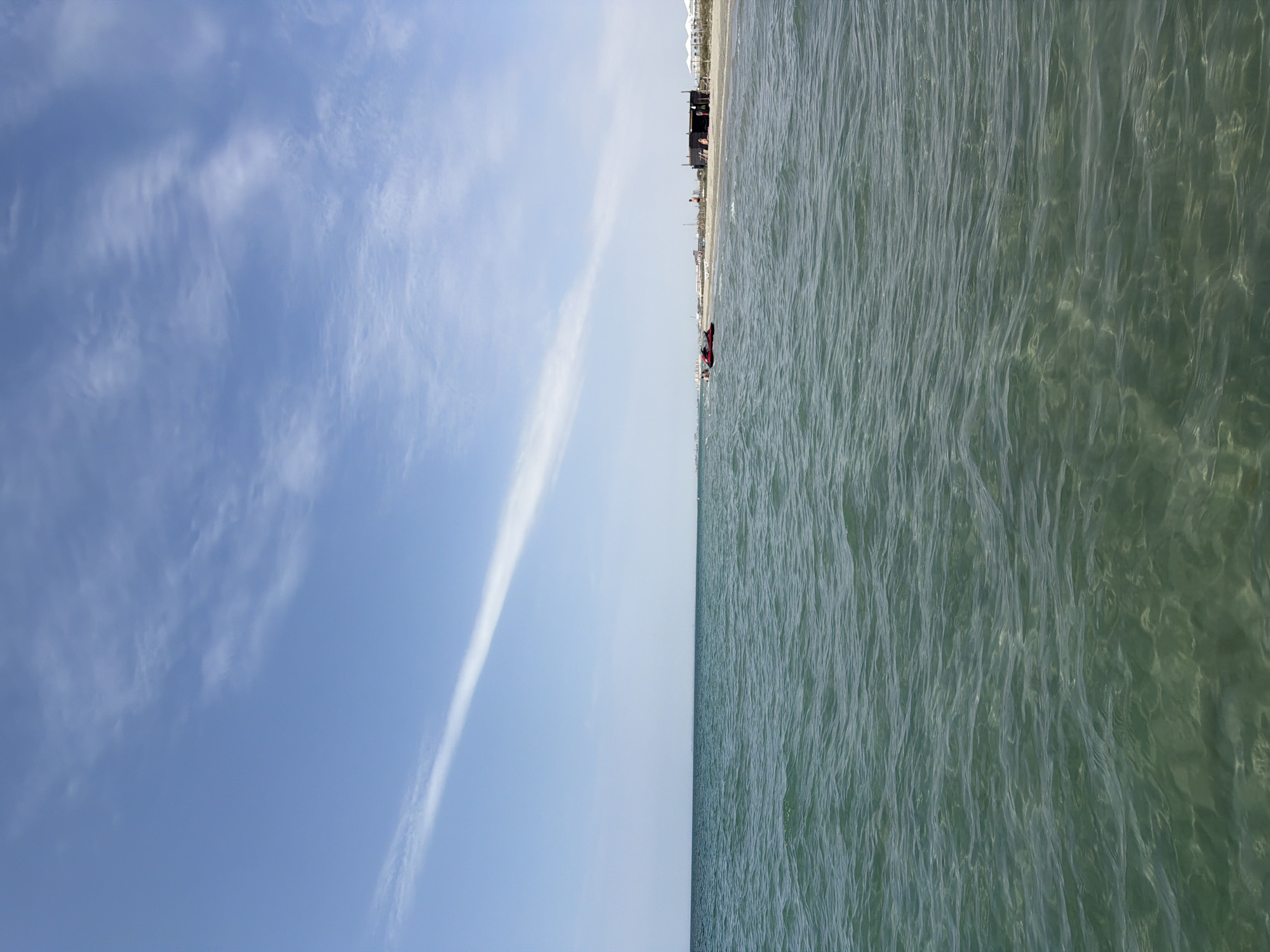 The image depicts a serene beach scene under a bright, partly cloudy sky. The foreground is dominated by the rippling turquoise water of the sea, which appears shallow enough to see the sandy bottom in places. A thin, wispy white cloud stretches diagonally across the upper portion of the sky, contrasting with the clear blue expanse.
In the middle ground, the sandy beach stretches along the horizon. Several structures are visible, including what appears to be a dark-colored beach hut or cabana with an open side, and several white umbrellas or awnings suggesting a resort or rented beach spots. A few people are scattered on the beach, appearing as small figures. Closer to the water, there are two red jet skis parked on the sand.
The overall atmosphere is one of a calm, sunny day at the beach, likely during daylight hours. The location is identified as being in the city of Unknown, Kazakhstan.