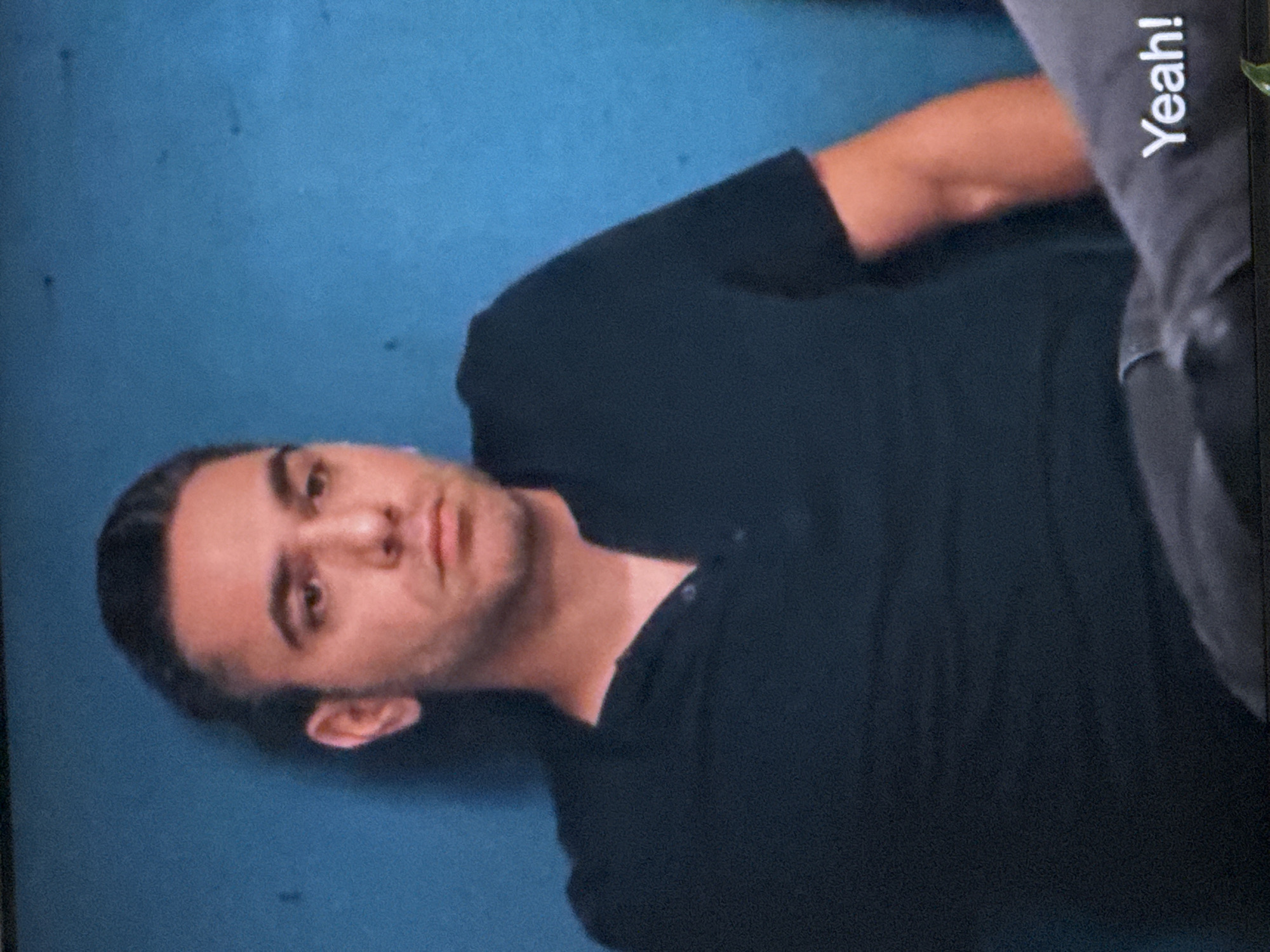 In this close-up shot of a man, he is leaning against a blue wall. He is wearing a black Henley shirt. His hair is dark and slicked back. His expression is neutral, and he is looking slightly off-camera. In the foreground, the word "Yeah!" is visible in white text on a grey surface, possibly a piece of clothing or furniture. The image is grainy, suggesting it may be a screenshot from a video. There is no discernible information about the location in Springboro, United States of America.