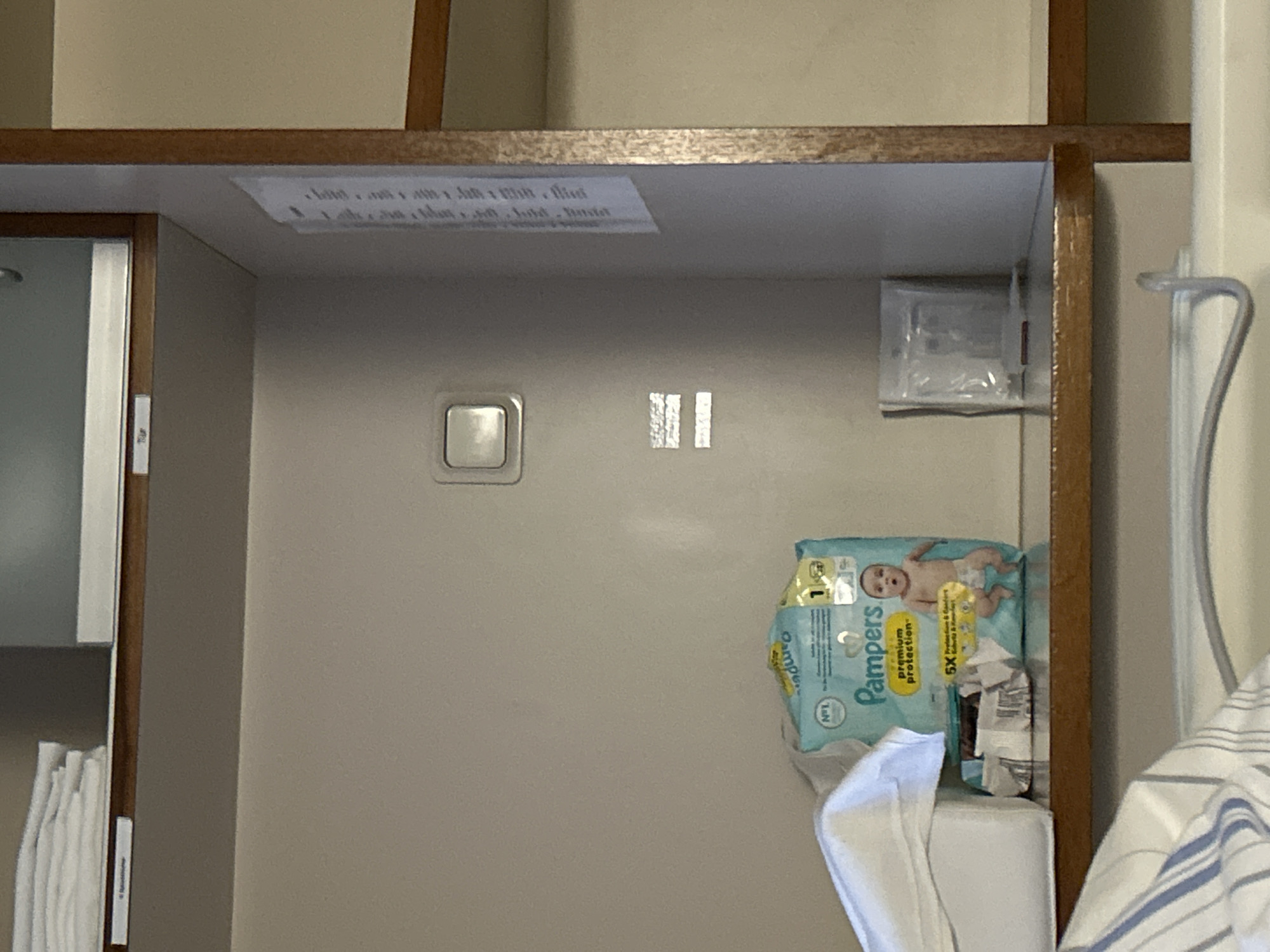 The image depicts a shelf within a room, possibly a hospital or care facility given the presence of Pampers diapers and other items. The shelf is made of wood and is part of a larger storage unit with neutral-colored walls. On the shelf, a pack of Pampers diapers is prominently displayed along with a white cloth item that could be a folded towel or baby blanket. To the right of the diapers, a small, clear plastic package containing what appears to be medical supplies is visible. In the background, a light switch is mounted on the wall. A document with vertical text is affixed to the right-hand side of the unit. The image is captured from a slightly elevated angle, focusing on the shelf and its contents.