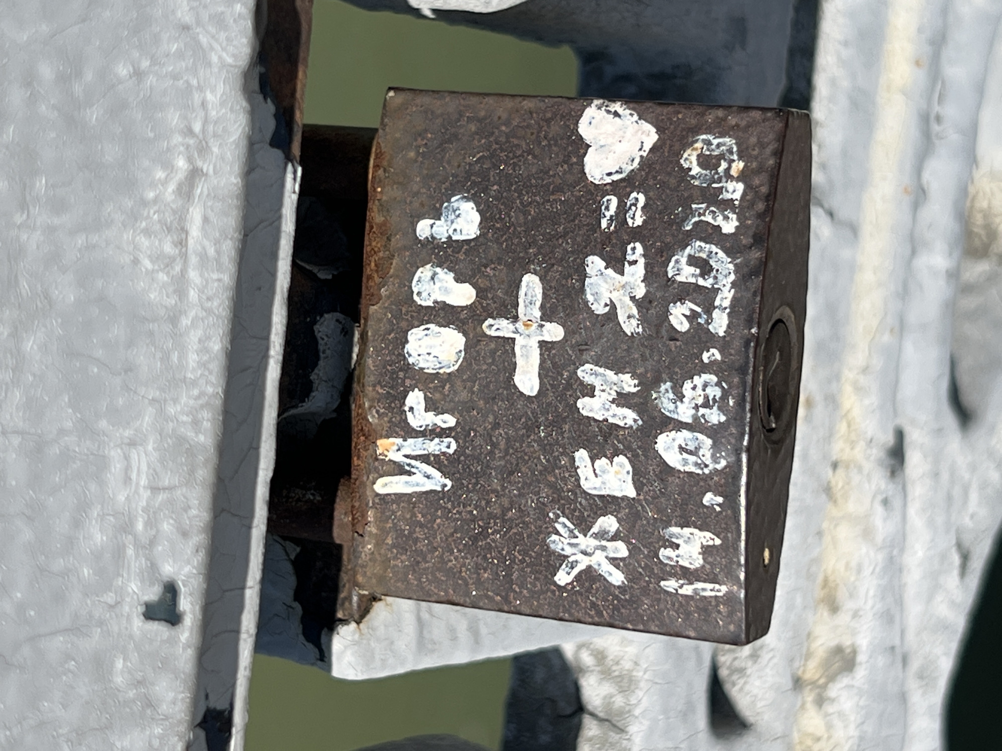 The image shows a close-up of a rusty padlock attached to a metal structure, possibly a bridge or fence. The padlock is inscribed with white paint or a similar substance. The inscription includes a heart symbol, the initials "V+," a cross symbol, the names "Irena" and "He," an asterisk symbol, and the date "14.06.2019." The metal structure appears to be painted white or light grey, and the background is blurry, suggesting a body of water or open space. This is located in Krasnodar, Russia.