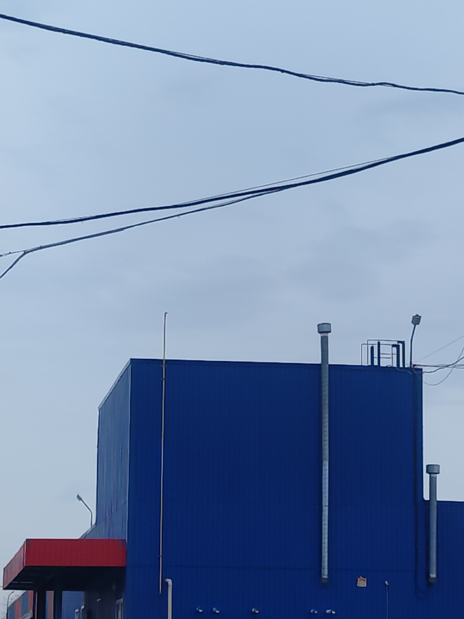 The image shows the exterior of a large, modern building located in Chaikovsky, Russia. The building has a boxy, industrial design, with blue metal paneling dominating the facade. A red overhang is visible, suggesting an entrance or loading area. Several pipes and vents protrude from the roof of the building, along with a small metal railing and a security light. Overhead, electrical wires stretch across the sky, which is overcast.