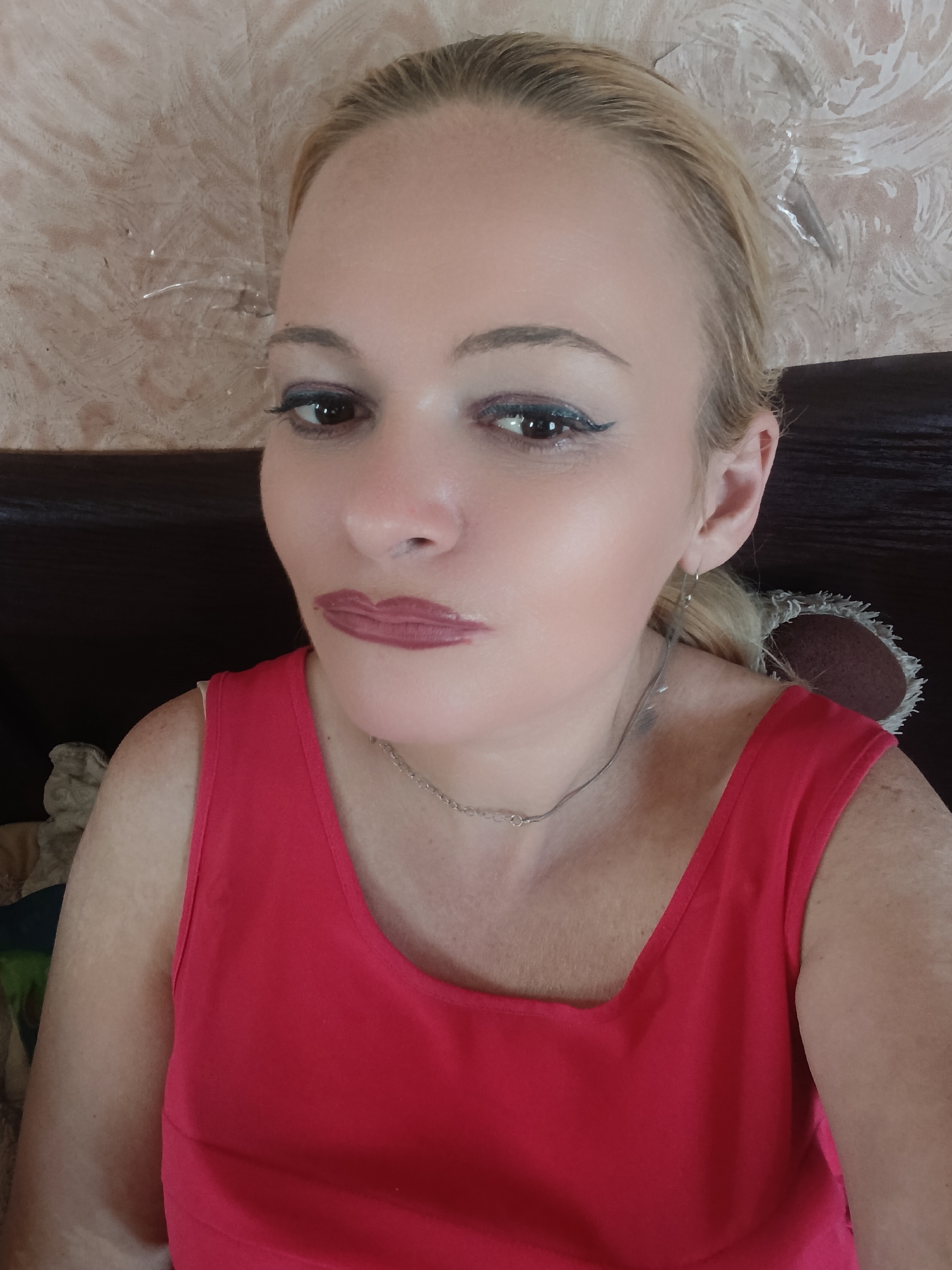 A woman with blonde hair pulled back is captured in a close-up, head-and-shoulders shot. She is wearing a bright red sleeveless top and a delicate silver chain necklace. Her makeup includes eyeliner and lipstick. Her gaze is directed slightly off-camera. The background consists of a patterned wallpaper and a dark wooden surface. The lighting suggests an indoor setting, possibly during the daytime, with no indication of specific weather conditions. The overall impression is a casual, personal photograph. There is no visible text in the image.