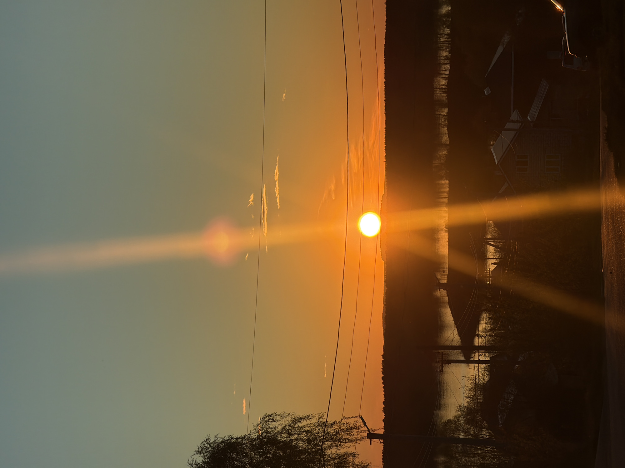 The image shows a sunset over a landscape in Chaikovsky, Russia. The sun is centered in the frame, with a bright glare emanating from it. Power lines cross the sky. A line of trees and vegetation is visible on the right side. The silhouettes of a building and streetlights are also visible. The left side of the image shows a gradient of colors, with blue transitioning into orange near the sun. In the bottom left corner, the top of a tree is visible.