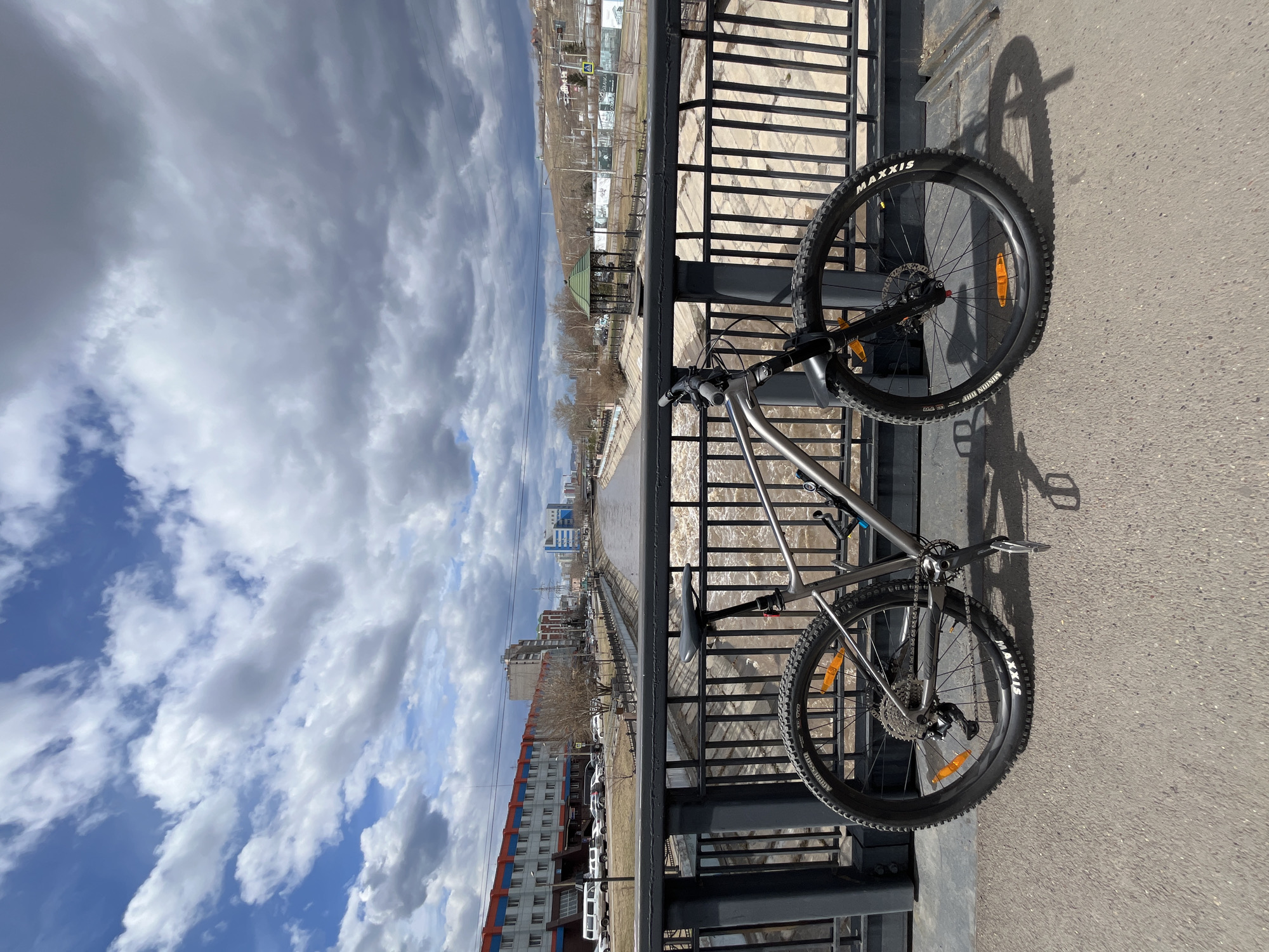 The image captures a mountain bike resting on a bridge under a partly cloudy sky, overlooking an urban landscape and a river. A silver-grey bicycle with prominent black "MAXXIS" branded tires, displaying "MINION DHF" and "AGRESSOR" models, leans against a dark, ornate metal bridge railing. The bike casts a distinct shadow on the asphalt surface.
Beyond the railing, a wide body of brownish, sediment-rich water flows, indicative of a river or canal, possibly influenced by snowmelt. The far bank reveals an urban environment with multi-story buildings, some featuring sections of orange-red and blue facades. Bare trees are scattered across the landscape, suggesting either late winter or early spring. Hills or higher ground are visible in the distant background.
The scene takes place during the daytime, likely late morning or early afternoon, given the bright illumination and the angle of the shadows. The weather is partly sunny with a mix of fluffy white and grey clouds against a blue sky. No people are visible, and the bike appears stationary, possibly paused during a ride or positioned for the photograph. The urban setting with a river and surrounding hills is consistent with locations like Krasnoyarsk, Russia.