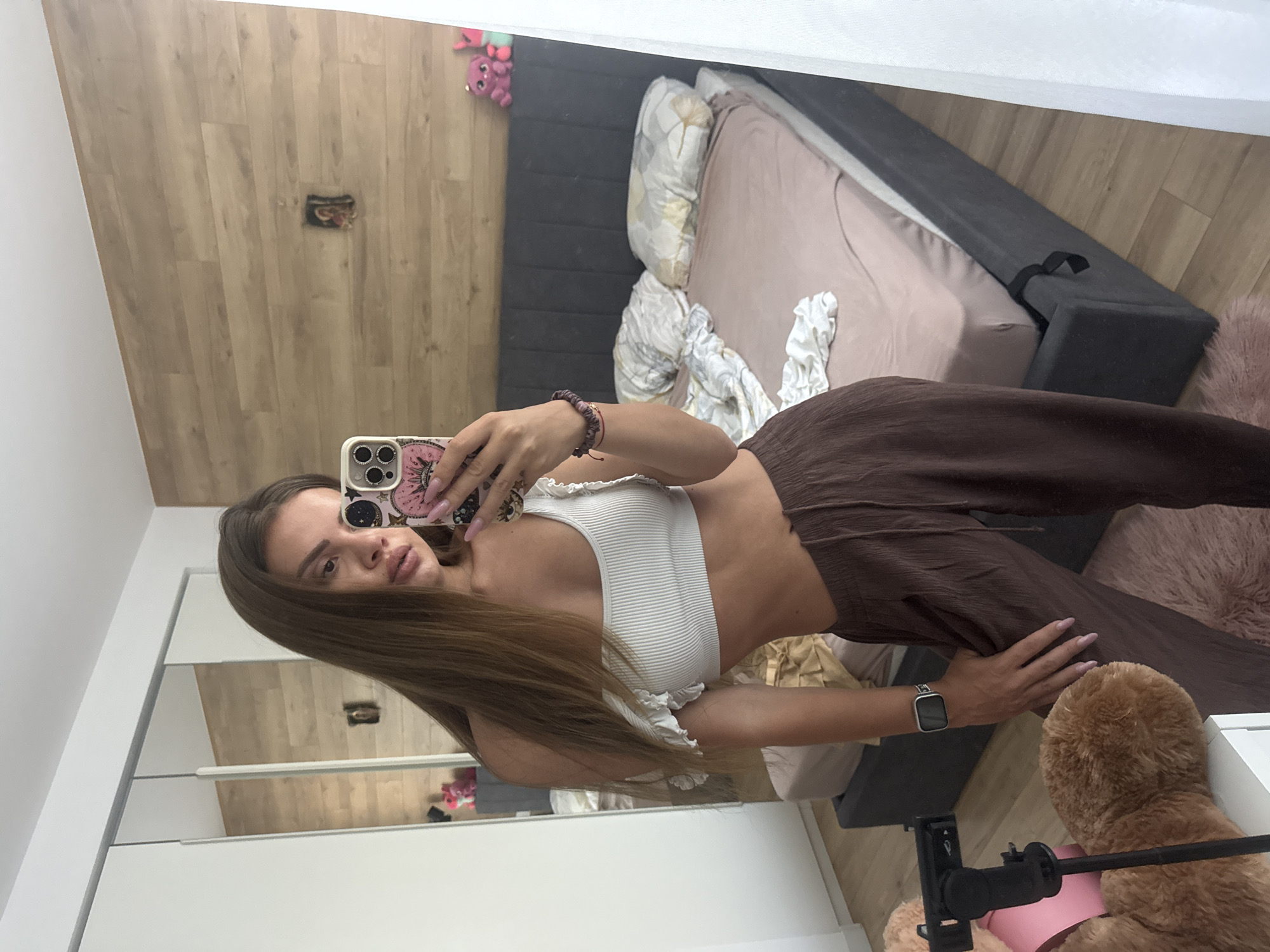 This is an indoor mirror selfie taken in what appears to be a bedroom, located in Plovdiv, Bulgaria. A woman, seemingly in her 20s or 30s with long, straight brown hair, is visible from the mid-thigh up, posing and holding a silver-cased smartphone with a pink patterned cover in her right hand to capture her reflection. She is wearing a white ribbed off-the-shoulder crop top with ruffled details on the sleeves and loose-fitting dark brown pleated pants with a visible drawstring waist.
On her left wrist, she wears a silver-framed smartwatch with a dark band. A multi-strand bracelet is visible on her right wrist. Her fingernails are painted a light pink. The room features a light wood laminate floor, with a small, shaggy pink rug positioned near her feet. To the immediate left, a portion of a large brown teddy bear is visible next to a black smartphone tripod stand.
Behind the woman, the reflection shows a queen-sized bed with a dark grey upholstered headboard and frame. The bed is covered with a light pink fitted sheet, and two pillows (one floral patterned, one white with a grey pattern) are visible at the head of the bed, along with a crumpled white blanket. The wall behind the bed is clad in light-colored horizontal wooden panels, featuring a small dark framed picture/icon mounted on it. Two small stuffed animals (one pink, one teal) are placed on the headboard to the right. To the far left, a white sliding-door wardrobe with mirrored panels reflects more of the room. A white curtain partially obscures the far right side of the image, suggesting a window.