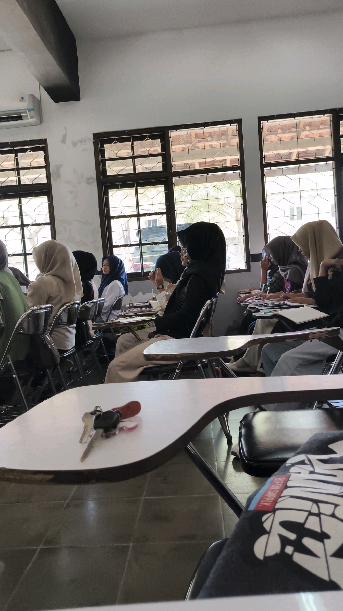 The image captures an indoor scene, likely a classroom, in Kartasura, Indonesia. The room features large windows with wooden frames, letting in natural light. Several individuals, predominantly women wearing hijabs, are seated at desks, seemingly engaged in an activity such as attending a lecture or studying. Some are looking towards the front of the room, while others appear to be writing or looking at their notes. The desks are simple, with attached writing surfaces. In the foreground, a set of keys with a red fob rests on a white desk. The floor is tiled. The overall impression is one of a typical educational setting.