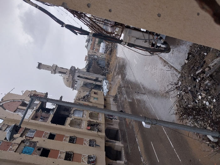 This image displays a scene of significant urban destruction in Khan Yunis, Palestinian Territory, under an overcast sky. A severely damaged multi-story beige building occupies the left side, featuring extensive structural compromise, visible interior spaces, and numerous broken windows. A grey street lamp post stands in the immediate foreground, slightly left of center. Behind the street lamp, a wet asphalt road with white lane markings recedes into the distance. In the mid-ground, a heavily damaged mosque with a distinctive dome and a partially collapsed minaret is visible amidst debris. To the right, a section of another beige building shows structural damage, with a utility box and bundled cables affixed to its exterior. Piles of concrete rubble and assorted debris are scattered across the foreground and along the wet street edges. The ground is consistently wet, suggesting recent rainfall or standing water. No individuals are discernibly present within the depicted area.