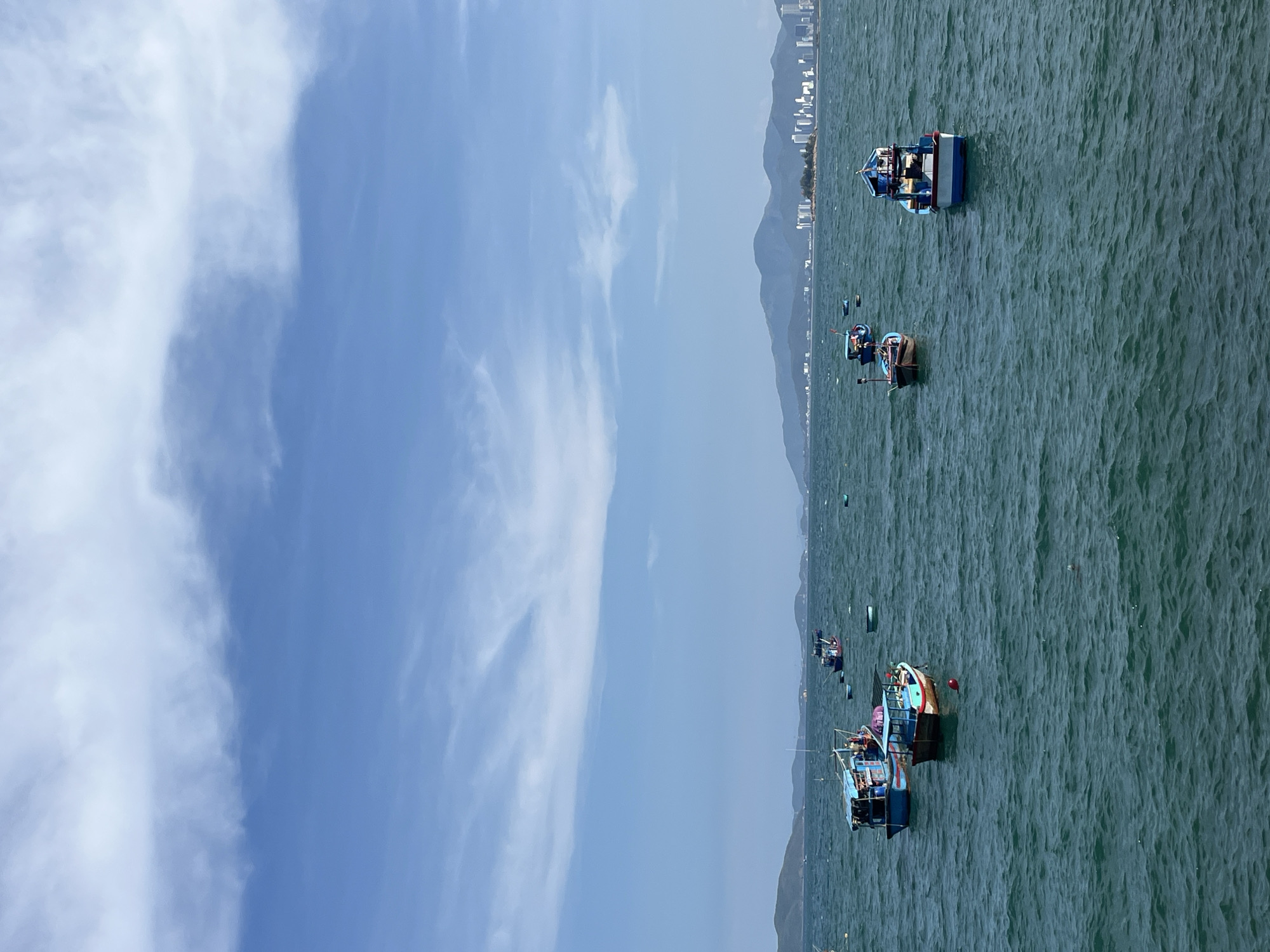 The image shows a coastal scene in Nha Trang, Vietnam, likely off the coast of Bac Nha Trang Ward. The sky is partially cloudy, with blue sky visible. The sea is a dark turquoise green, with visible wave patterns. Several fishing boats are scattered across the water, varying in size and color. Some boats are closer to the viewer, showing more details like structures and gear on board. A mountain range is visible along the coastline, with a stretch of buildings suggesting urban development. The photograph is taken from an elevated viewpoint.