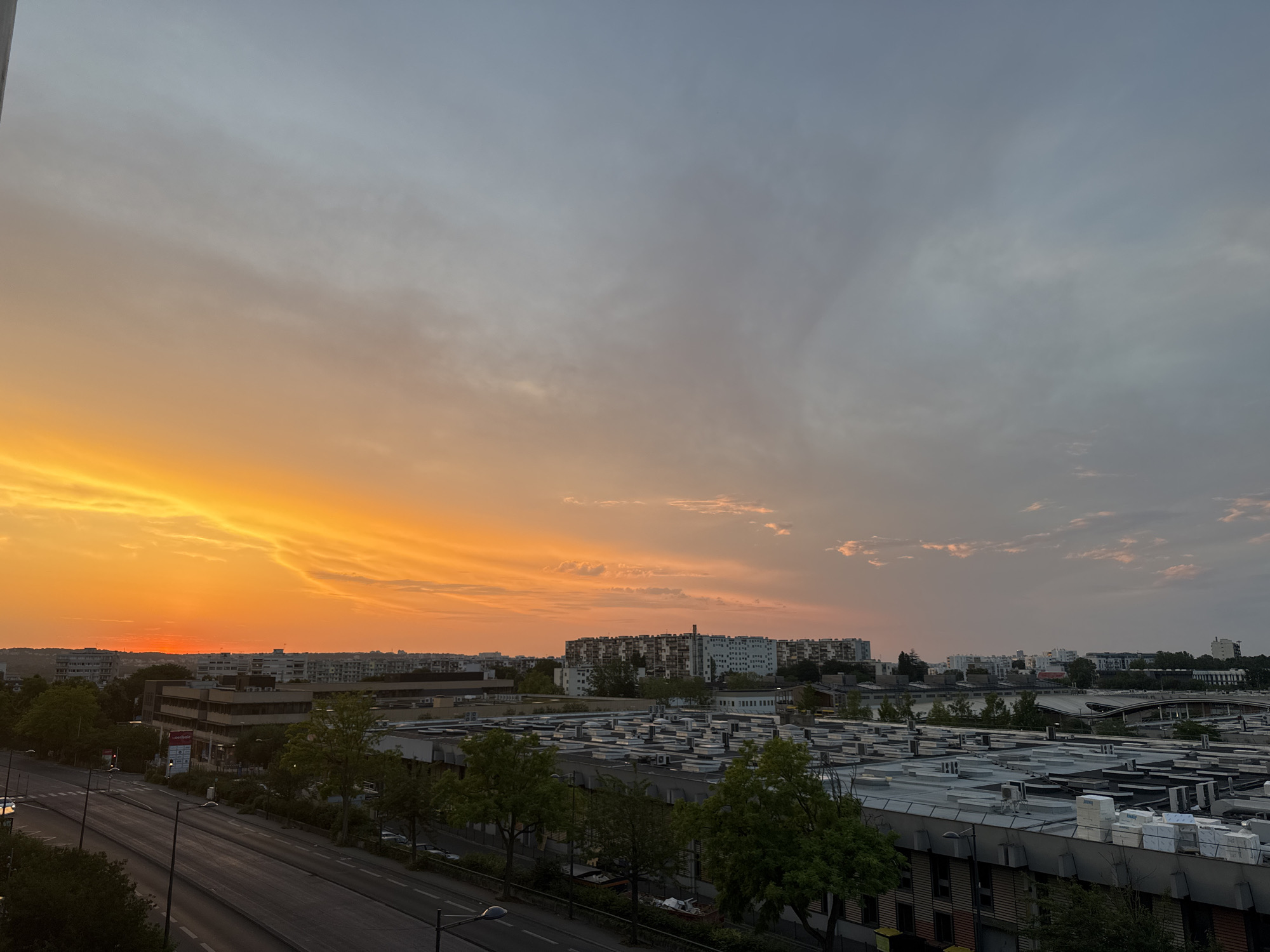 This is a high-angle, wide shot of a city skyline at sunset. The sky is a gradient of warm orange and yellow near the horizon, transitioning to a soft gray and blue at the top. Wispy clouds are scattered throughout the sky. In the foreground, a multi-lane road runs diagonally from the bottom left to the center of the image. On either side of the road are sidewalks and trees. Beyond the road, there is a dense urban landscape with various buildings. A prominent feature is a large, flat-roofed industrial building with many air conditioning units and what appear to be stacks of materials on its roof. Further back, a cluster of taller residential buildings can be seen, some with balconies. The overall impression is of a moderately dense urban area during a colorful sunset, with a mix of commercial and residential structures. There are no people visible in the frame.