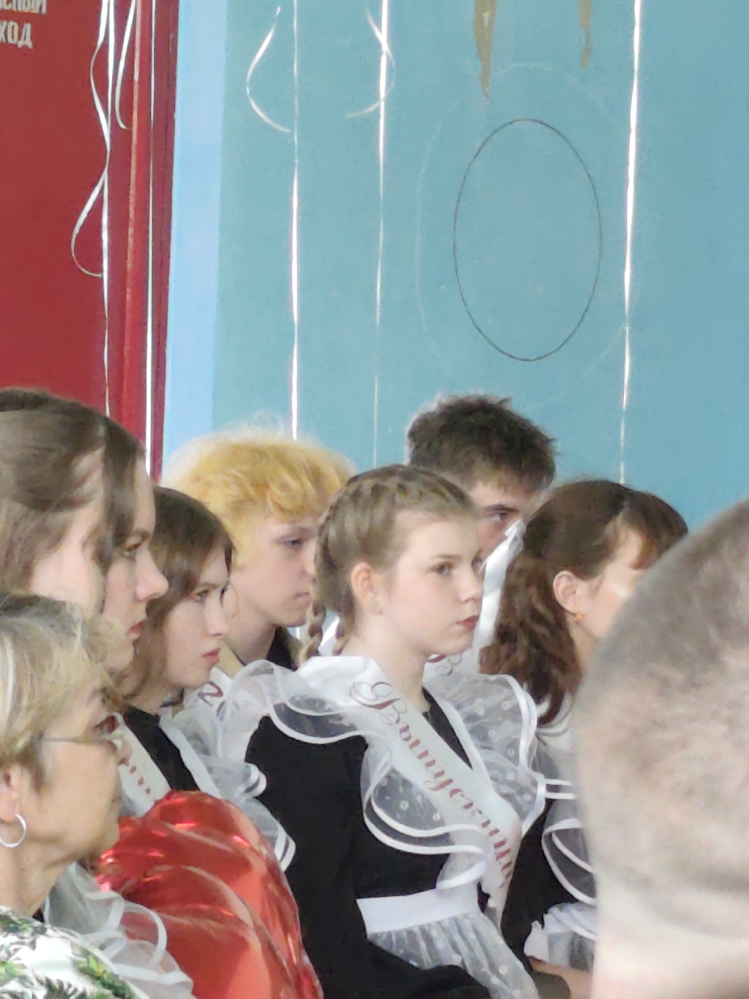 The image depicts a group of people, seemingly gathered indoors. Several individuals are identifiable as young women in school uniforms with decorative sashes, which may suggest a graduation or celebratory event. One woman is wearing glasses and earrings, and she is situated near a partially visible red object that could be a balloon.
The location cannot be verified as Gremyachinsk, Russia based on the visual information provided in the image. There are wall features that suggest an interior space like a school assembly hall.