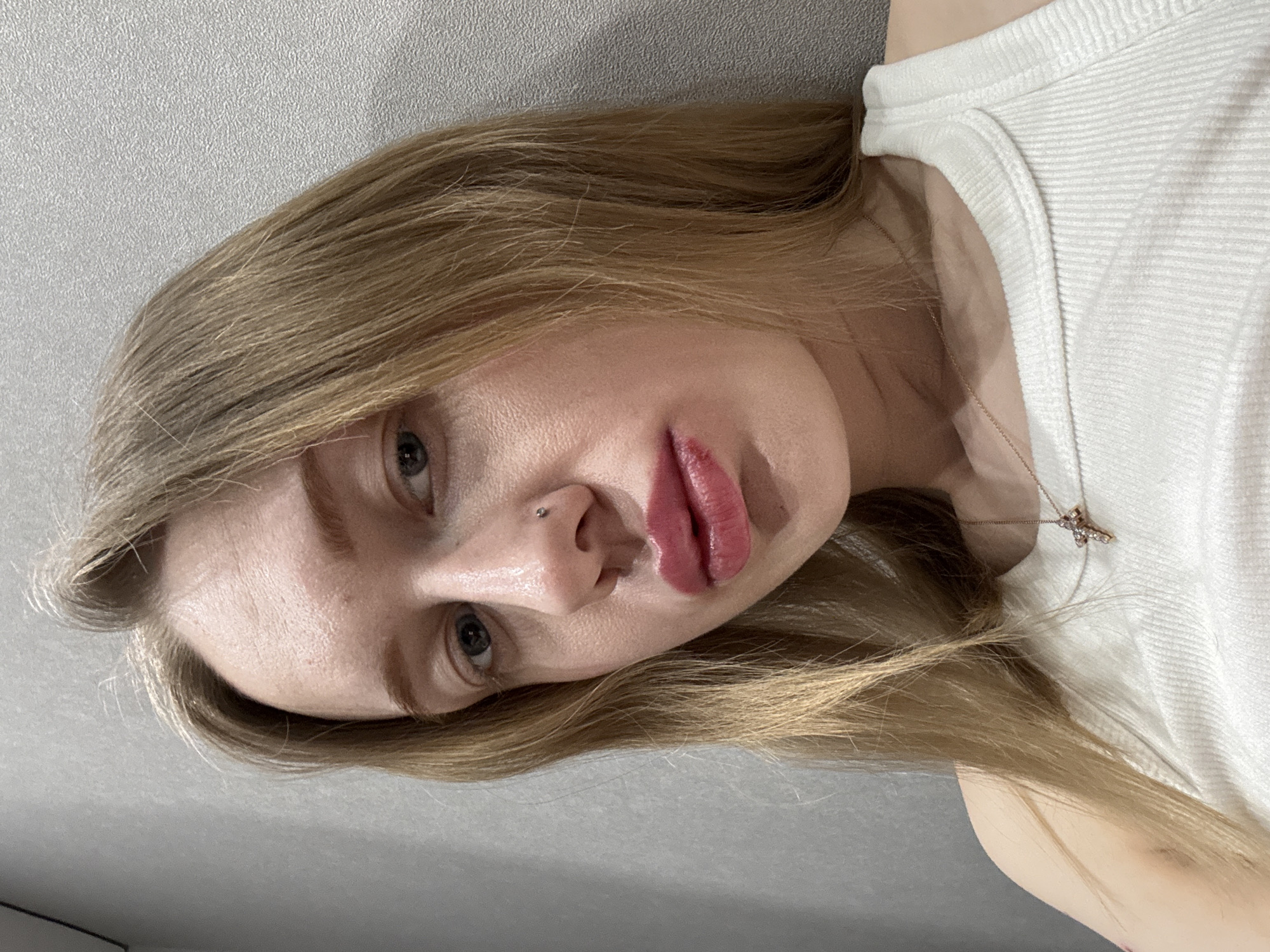 A young adult female is depicted in a close-up, chest-up portrait, positioned against a plain, light grey textured wall. She possesses fair skin, long straight blonde hair, and blue-grey eyes looking directly at the camera. A small stud piercing is present on her left nostril. Her lips are full and coated with a glossy pink-red substance. She is wearing a white ribbed tank top and a thin, gold-colored chain necklace with a small, stone-embellished cross pendant. The specific location within the city of Rostov-on-Don, Russia, is not ascertainable from the visual information provided in the image.
