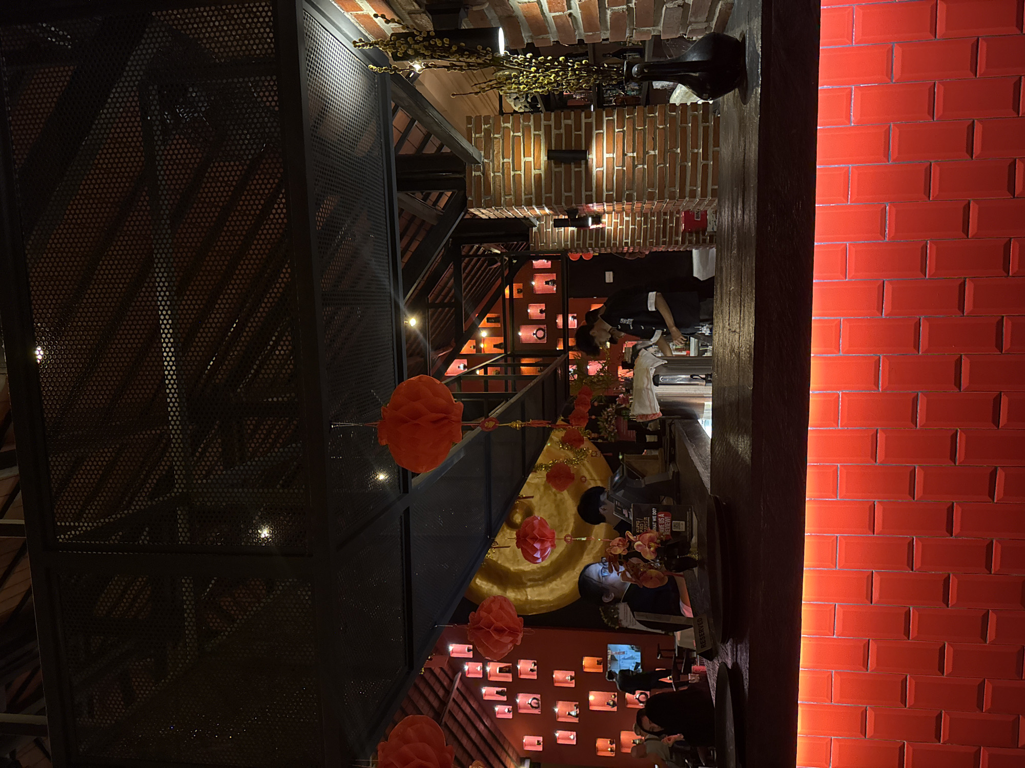 The image depicts the interior of a restaurant in Kuta, Indonesia, characterized by a warm and festive ambiance, likely for a celebration. The decor features numerous red paper lanterns, a dominant color scheme of red and orange hues, and exposed brick walls, suggesting an East Asian or Asian fusion theme.
At the foreground, a dark wooden bar top is visible, with bright red tiled walls beneath it, illuminated from behind. Behind the bar, several individuals are present. To the left, a woman wearing glasses is looking at her phone, seated at the bar. Further back, a man in a black uniform appears to be working behind the bar. Another person in a similar uniform is also visible.
The background reveals a tiered shelving unit against a red wall, displaying various decorative items, possibly vases or pottery, each illuminated by a soft light. A large, round golden object, possibly a gong, hangs on the wall in the middle ground. The ceiling is industrial with visible metal beams and a perforated metal enclosure. Decorative yellow branches in a tall black vase are placed on the right side of the bar, adding to the overall decor.
The lighting is dim and atmospheric, with a focus on the red and orange tones, and highlights from the shelves and bar. The scene appears to be an evening setting.