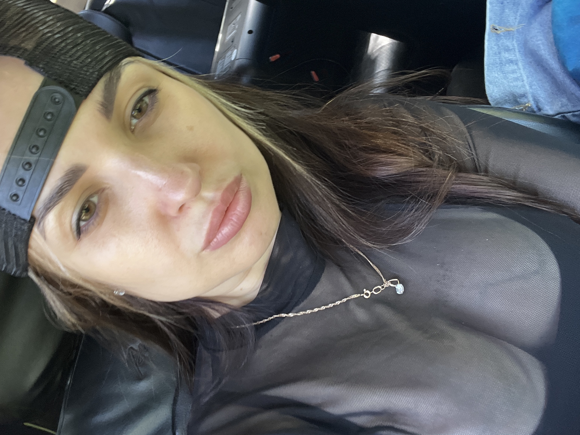 A close-up portrait shows a woman wearing a black trucker hat and a sheer black top with a gold necklace. Her hair is dark brown with blonde highlights. She is likely seated in a car due to the presence of car interior elements in the background. The location is unknown.
