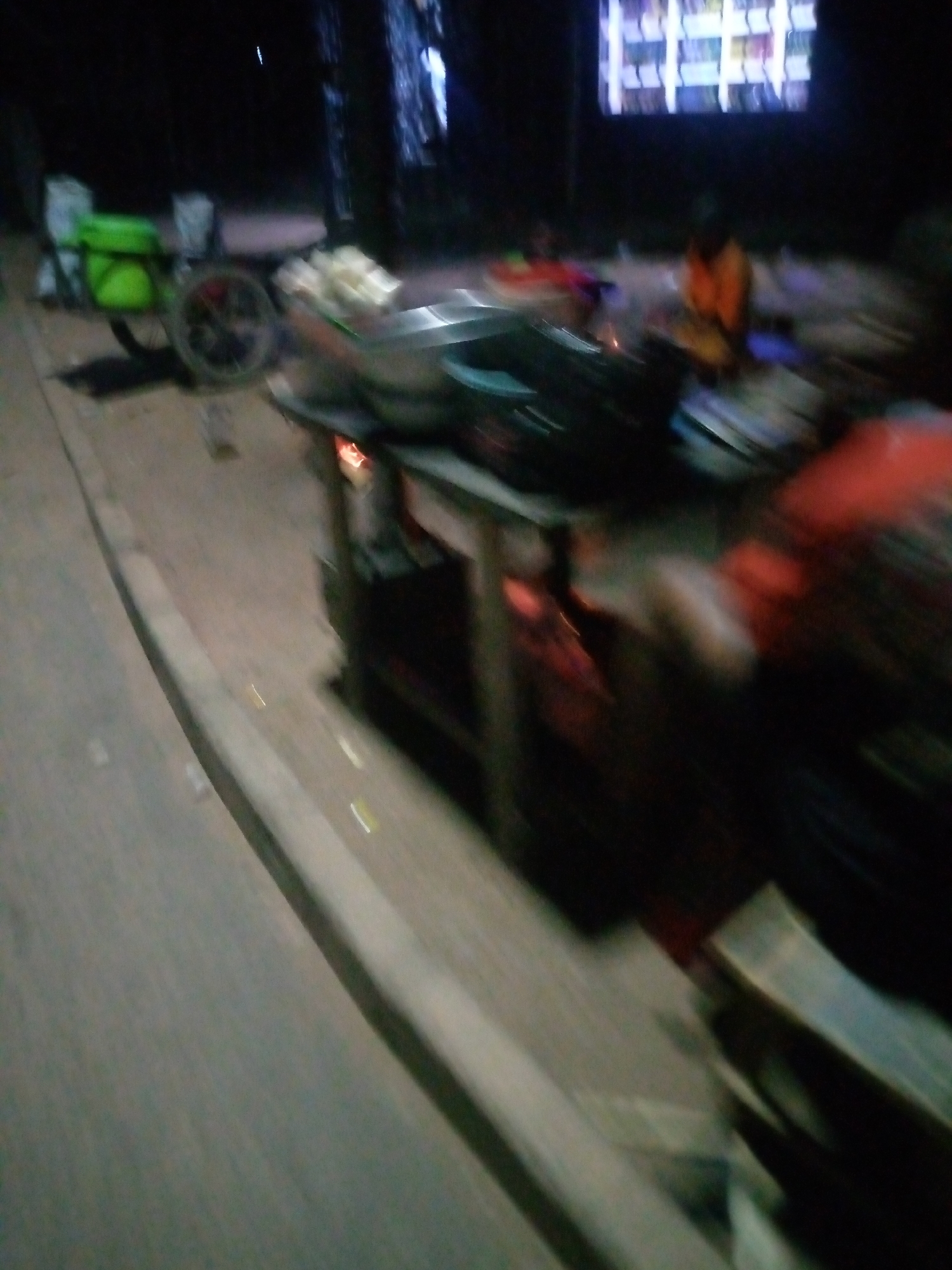 The image depicts a nighttime scene in the city of Jalingo, Nigeria. In the foreground, a street vendor's stall is visible, with various items arranged on shelves and a fire burning beneath. A cart with a green container and a wheel is parked nearby, suggesting it's used for transporting goods. In the background, a large screen displays a colorful grid of images, possibly an advertisement or display. A person in an orange garment is seated at the stall, and another individual in a red garment is partially visible. The environment appears to be an outdoor urban setting, with the street illuminated by artificial light. The overall impression is one of a bustling street market at night.