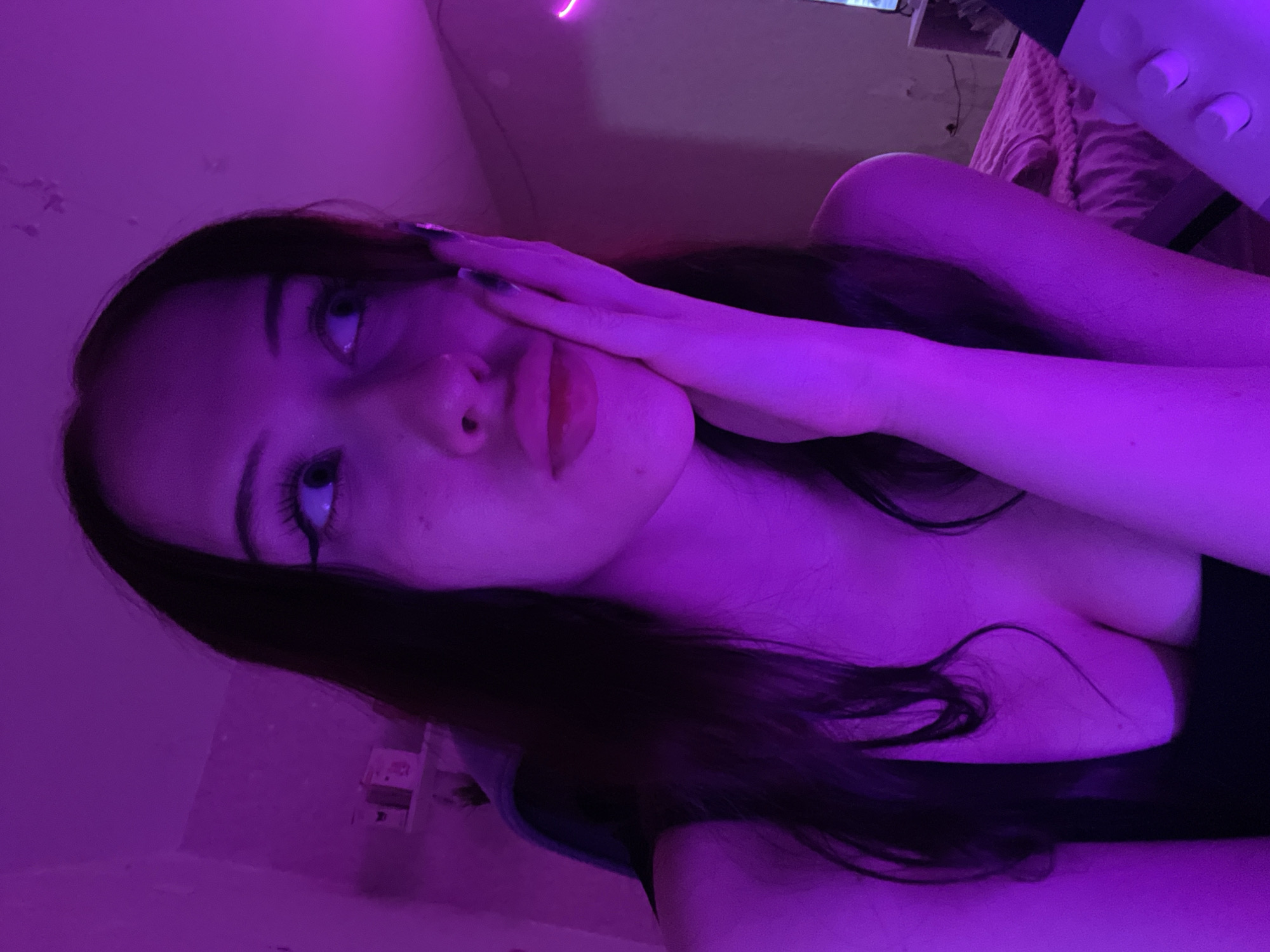 A young woman with dark hair and heavy eyeliner looks to her right, resting her chin on her hand. She is illuminated by purple light, which casts a neon glow on her skin and the surrounding room. Her expression is pensive, with slightly parted lips. She is wearing a dark, low-cut top. The background is dimly lit, showing hints of a wall and furniture, with a pink neon light visible on the right side. There is no indication of her location in Votkinsk, Russia.