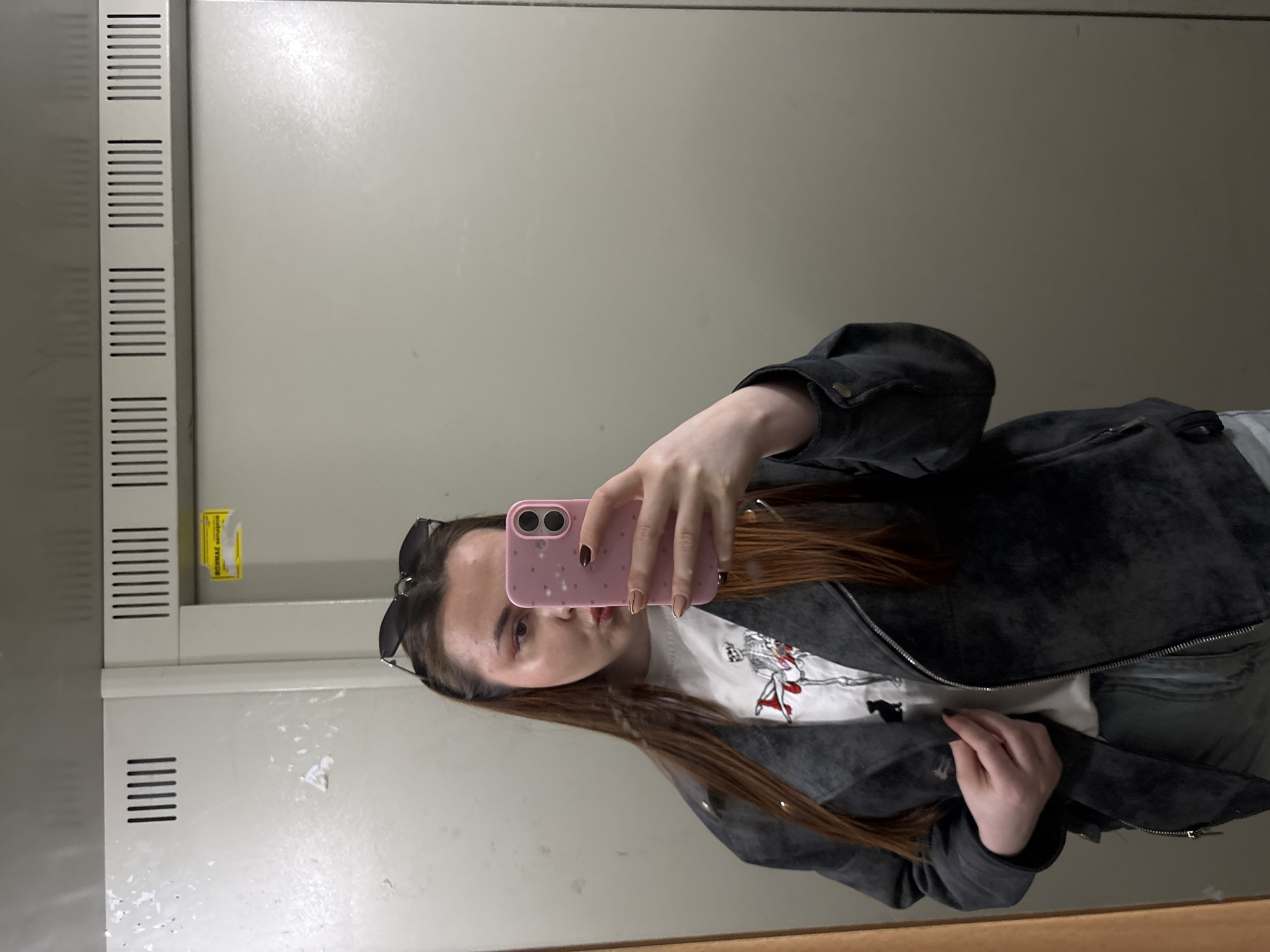 A young woman takes a selfie in a mirror. She is wearing a white t-shirt with a graphic print, a dark gray suede jacket, and has sunglasses perched on her head. She holds a pink phone with both hands, partially obscuring her face. The background appears to be an industrial or institutional setting with gray cabinets featuring ventilation slots. No information within the image can be used to identify Izhevsk, Russia.