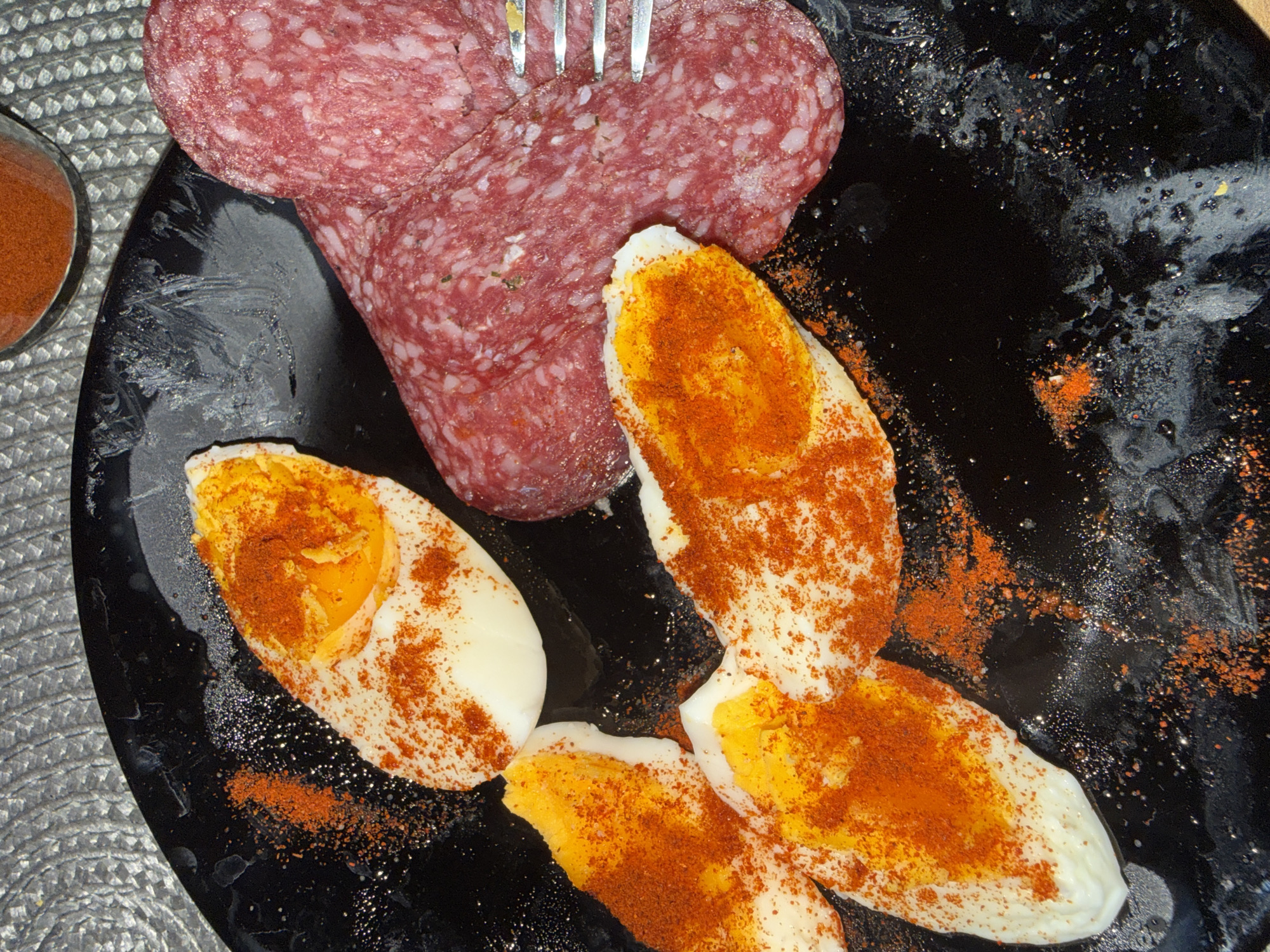 An overhead shot captures a simple, savory meal arranged on a glossy black plate, set against a textured, silvery-grey woven placemat. The main subjects are several thick slices of salami and multiple halved hard-boiled eggs, both liberally dusted with a vibrant red spice, likely paprika.
In the upper center of the plate, thick slices of salami display their characteristic marbled texture of lean meat and white fat. A silver fork is seen piercing one of these slices, suggesting the meal is either being prepared or is actively being consumed.
Below and to the right of the salami, several hard-boiled eggs are cut in half lengthwise, revealing their firm, bright yellow yolks and white albumen. These egg halves are generously covered with the same rich red spice, which also scatters across the surface of the black plate, creating a dynamic visual effect.
A small, clear glass container, partially visible on the left side of the frame, holds more of the red spice, indicating it's an intentional seasoning for the dish. The black plate itself shows a reflective, somewhat mottled surface with highlights and smudges, possibly from oil or moisture. The overall lighting is bright, emphasizing the colors and textures of the food. No people are visible, and there is no discernible text within the image. The scene presents a straightforward, ready-to-eat meal.