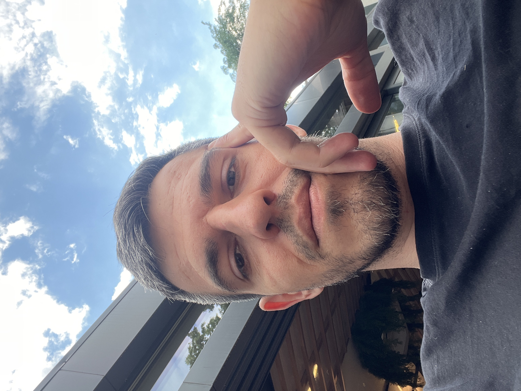 A close-up, low-angle selfie of a man with short, graying hair and stubble is taken outdoors, likely in Sofia, Bulgaria. He is wearing a dark t-shirt and has his right hand resting on his forehead. In the background, a modern building with reflective windows and a wooden ceiling structure is visible, along with greenery and a cloudy blue sky. The lighting suggests it is daytime.
