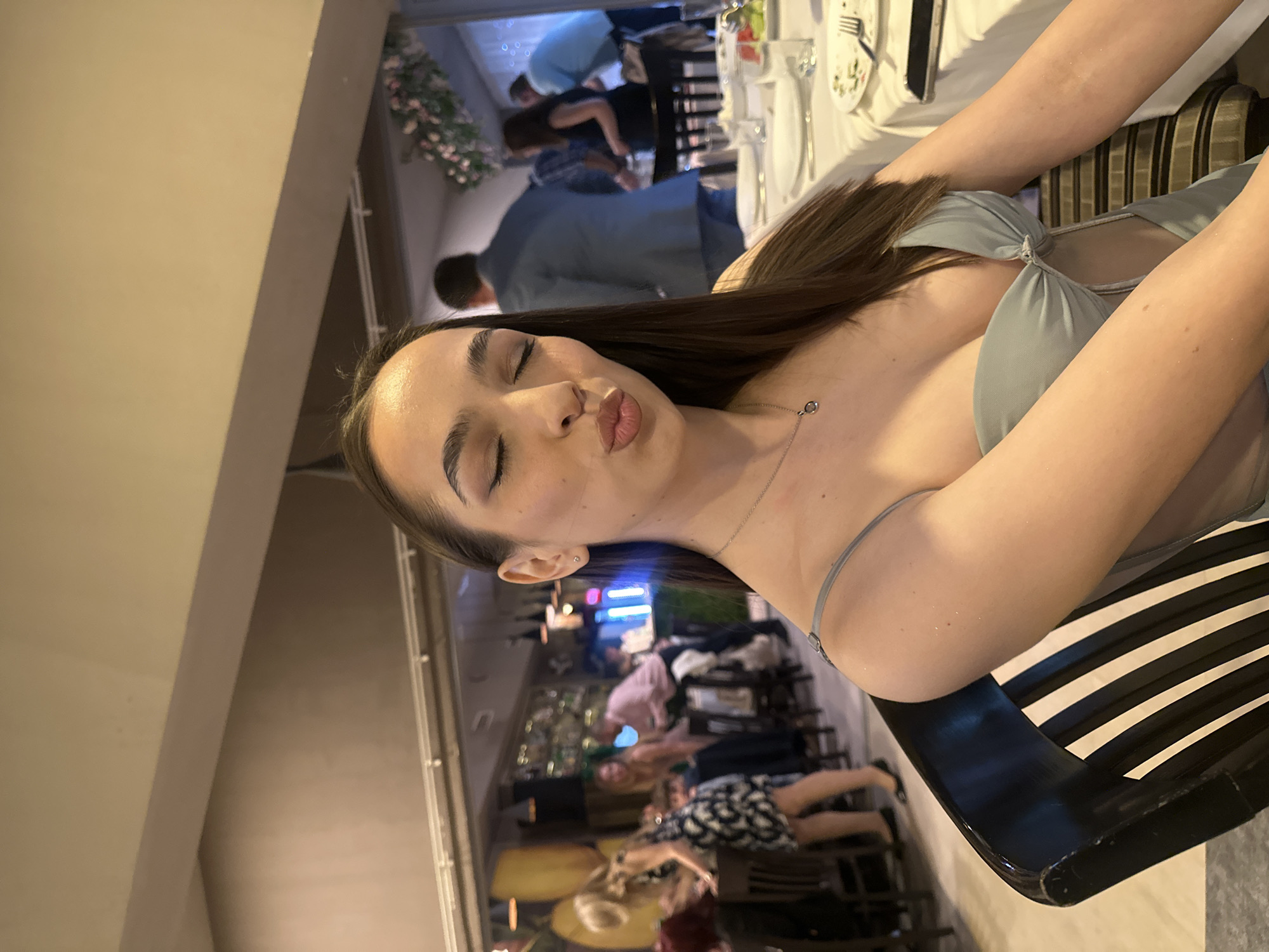A medium shot captures a young woman indoors, likely at an event in Sofia, Bulgaria given the likely location of the image. She is seated at a table, making a kissing face at the camera. Her eyes are closed. She has dark hair and wears a light-colored dress with thin straps. A delicate silver necklace adorns her neck.
Behind her, a blurred scene reveals other people at tables, suggesting a restaurant or gathering. Some individuals are walking around. The table in front of her has plates, glasses, and silverware, indicating a formal meal setting. A glimpse of what might be an outdoor area is visible through a window in the background. The chair she is sitting on has black and white stripes.