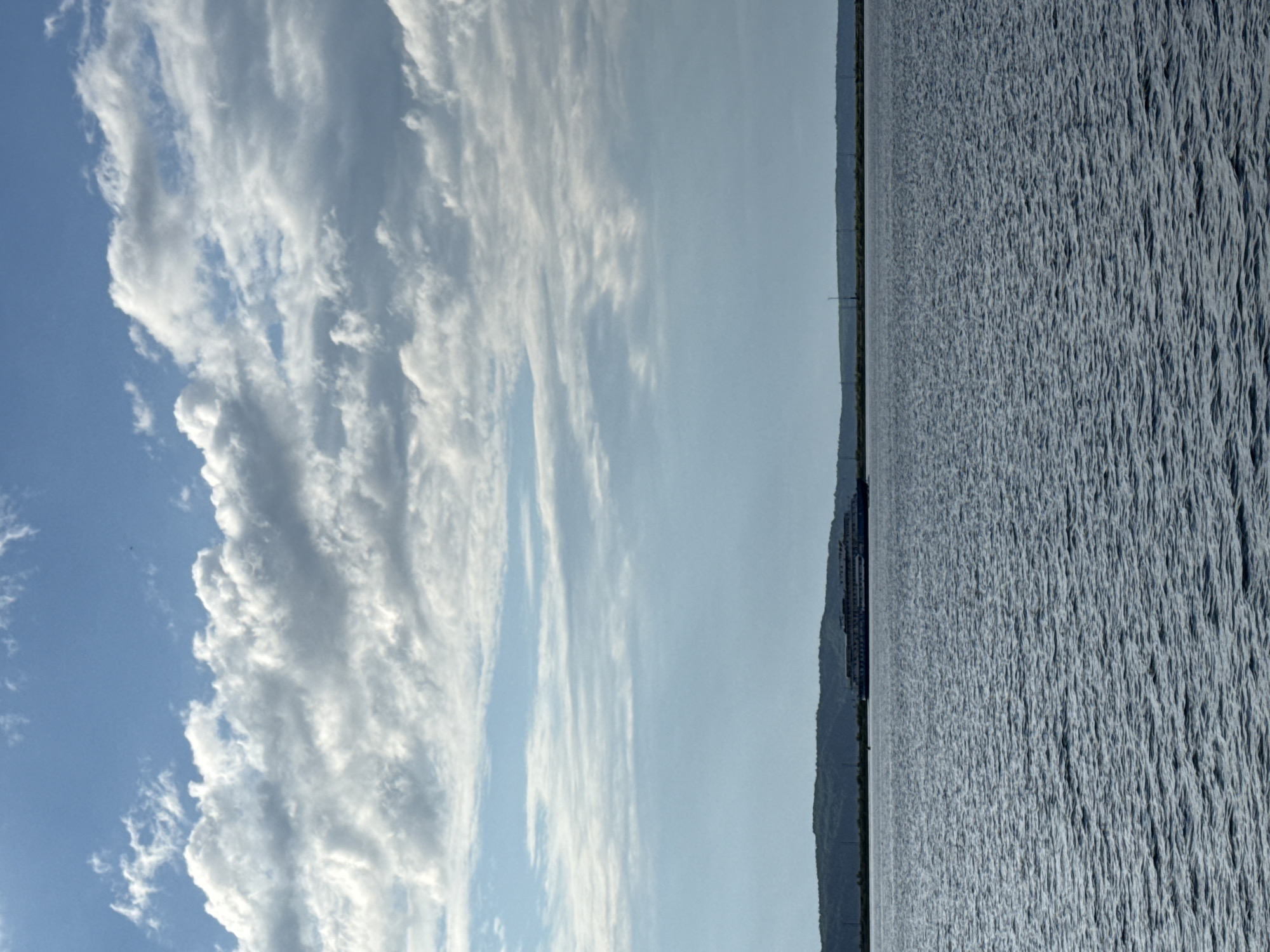 The image shows a vast, rippling body of water under a bright blue sky filled with fluffy white clouds. The water occupies the right side of the frame, its surface textured with numerous small waves. On the left side, the sky transitions from a clear, pale blue to a more dramatic expanse of clouds. A dark, elongated object, possibly a ship or a long building, is visible on the horizon, situated along a distant, tree-covered shoreline. The overall impression is one of a serene, natural landscape, likely captured on a clear day, perhaps in the late afternoon given the gentle lighting. There is no visible text in the image. The scene evokes a sense of tranquility and openness.