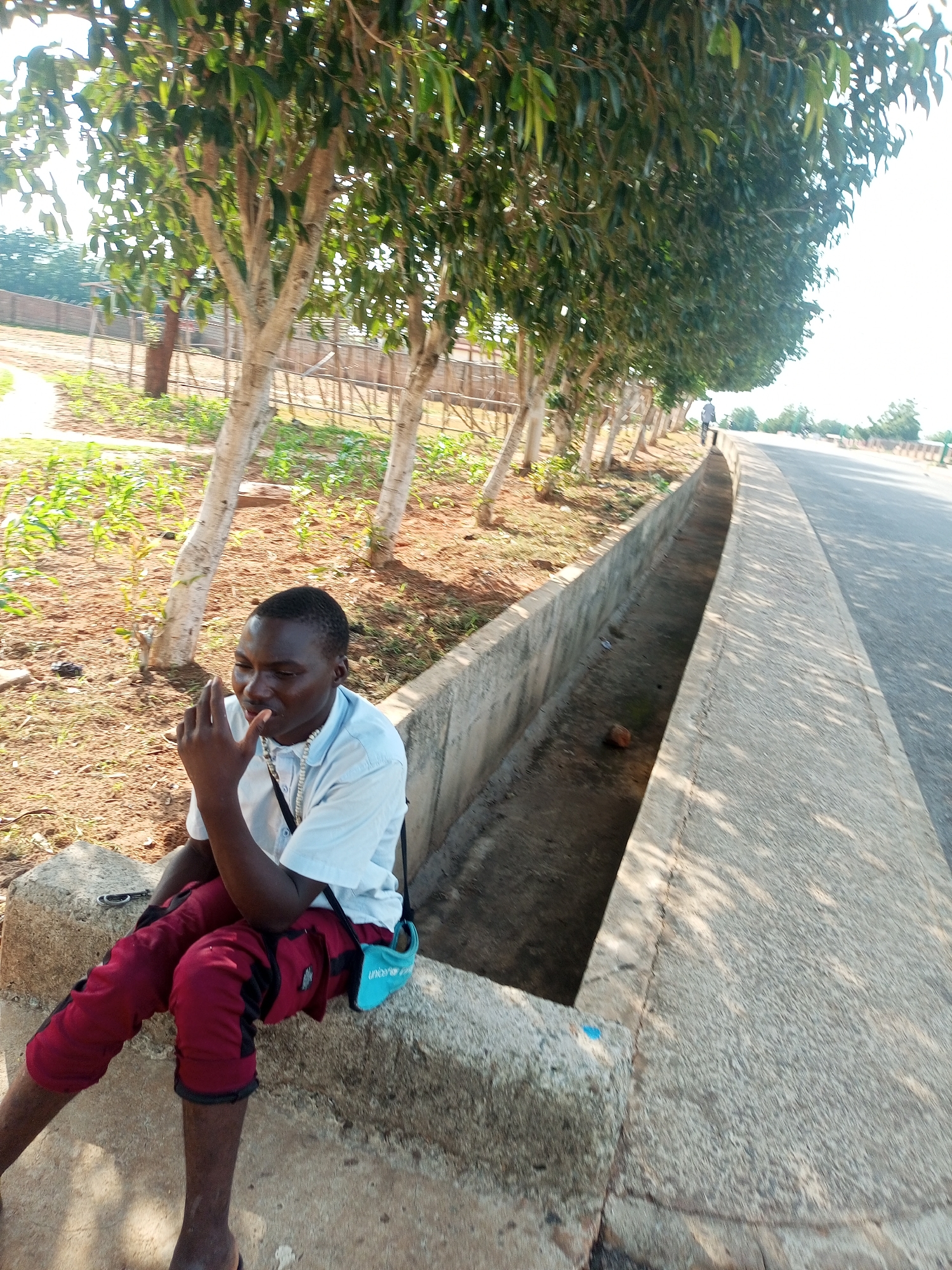 A young Black male is seated on a rough-hewn concrete ledge, his body angled to the right. He wears a light blue short-sleeved collared shirt and dark red cropped pants with black accents. A light blue pouch is attached to his right hip. He is holding a cigarette to his mouth with his right hand, fingers touching his lips. The setting appears to be an urban or semi-urban environment in Tempure, Nigeria, with a paved road to the right and a concrete drainage ditch running alongside it. A line of trees with light-colored bark and green foliage borders the scene on the left. The ground is covered in reddish-brown soil and sparse vegetation. In the background, a wooden fence and a few distant trees are visible under a bright, possibly overcast sky.