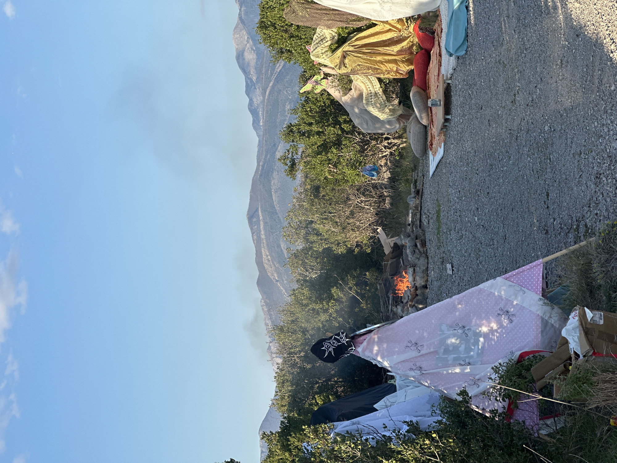 The image shows a campsite in Glavaticici, Montenegro, set against a backdrop of mountains and a clear blue sky. A fire is burning, surrounded by trees and vegetation. Assorted items are scattered around the campsite, including clothing, blankets, cardboard boxes, and other personal belongings. A pink blanket with a skull pattern is prominent near the fire. A gravel road borders the campsite. There are no people visible in the image.