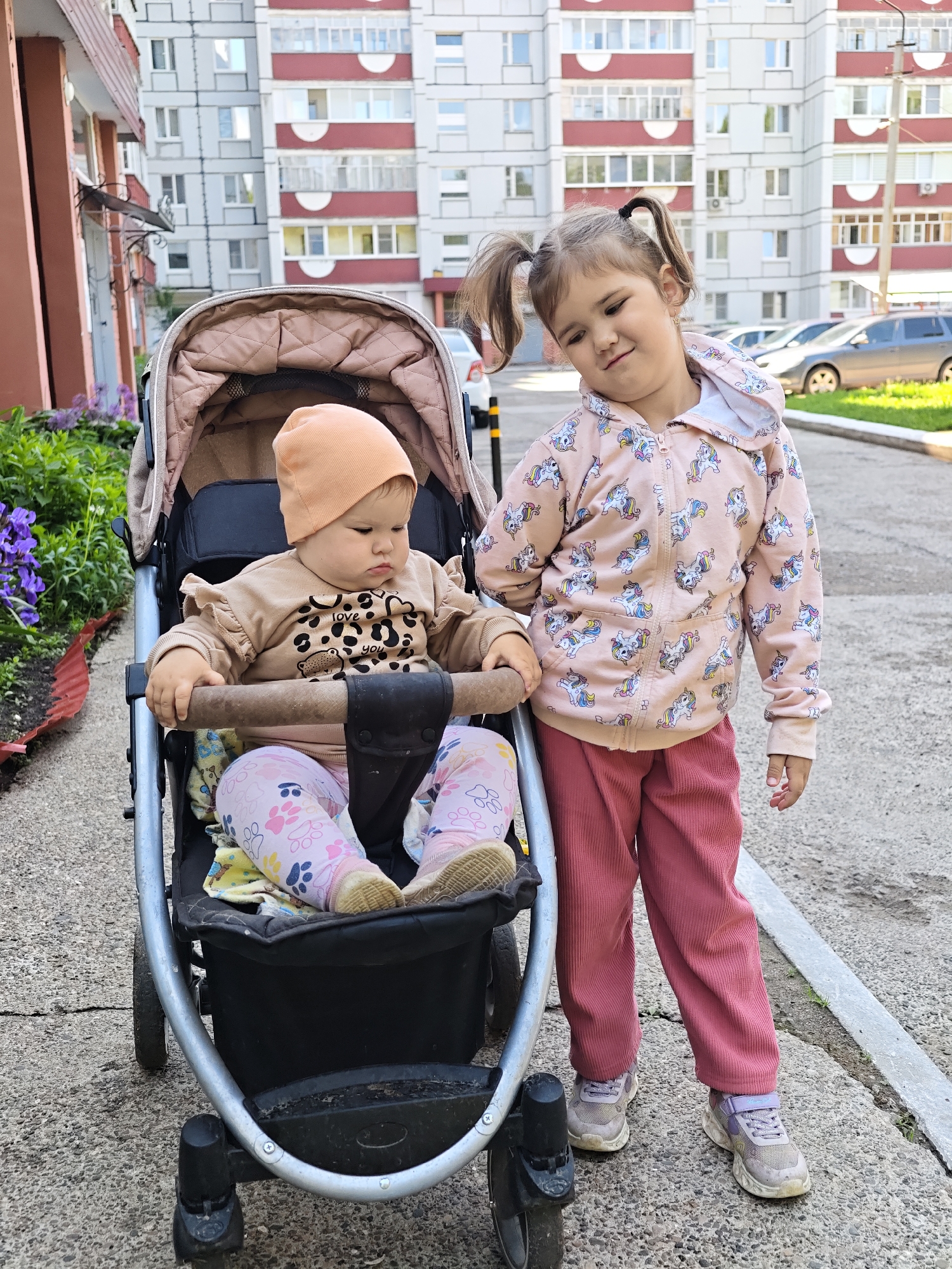 A daytime outdoor scene features two young girls in what appears to be a residential area in Chaikovsky, Russia. In the foreground, a baby or toddler sits in a beige and black stroller, facing slightly to the right. This younger child wears an orange beanie, a light brown long-sleeved shirt that reads "love you" with an animal print, and pink leggings with paw print patterns. The child has a somewhat serious or pouty expression.
Standing to the right of the stroller is an older girl, likely a preschooler, looking towards the camera with a playful, slightly mischievous smile and her head tilted. She has her hair styled in two pigtails. Her attire consists of a pink hooded zip-up jacket covered in unicorn patterns, dark pink corduroy pants, and grey and purple sneakers.
The background shows a large, multi-story apartment building with a light facade and red horizontal accents, consistent with typical residential architecture in the region. Several cars are parked in the distance behind the girls. To the left, there is a small patch of greenery with purple flowering plants. The ground is paved, showing some cracks. The scene is well-lit by natural light, indicating it is likely late morning or afternoon on a clear or slightly overcast day.