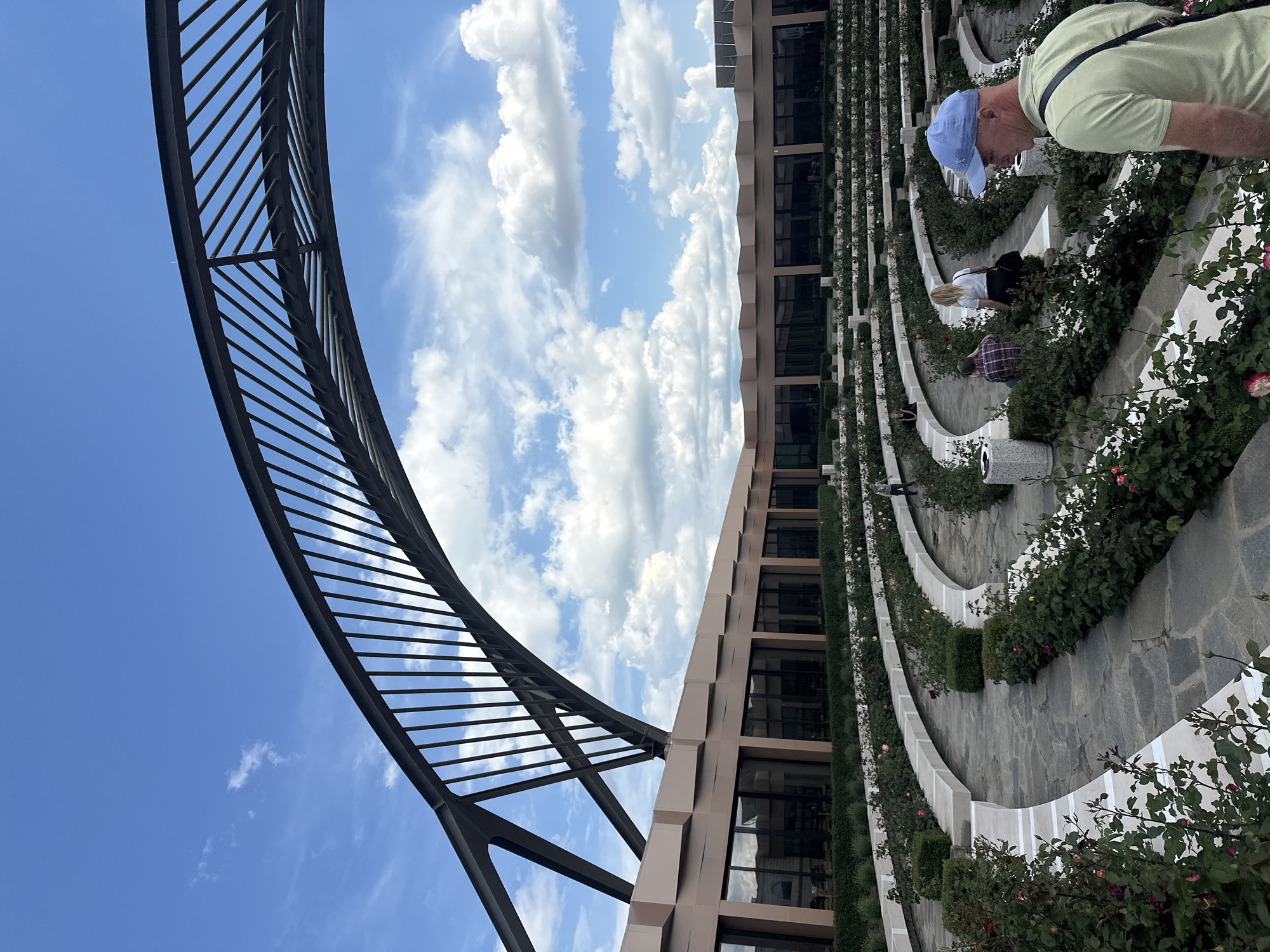 The image shows a tiered garden landscape with rows of rose bushes and stone pathways. People are present, appearing to be walking or observing the garden. A modern building facade with large windows stands to the side of the garden. A black metal structure with a curved design is visible against a backdrop of a blue sky with fluffy white clouds. The location is possibly Skobelevo, Bulgaria, as indicated by the prompt.