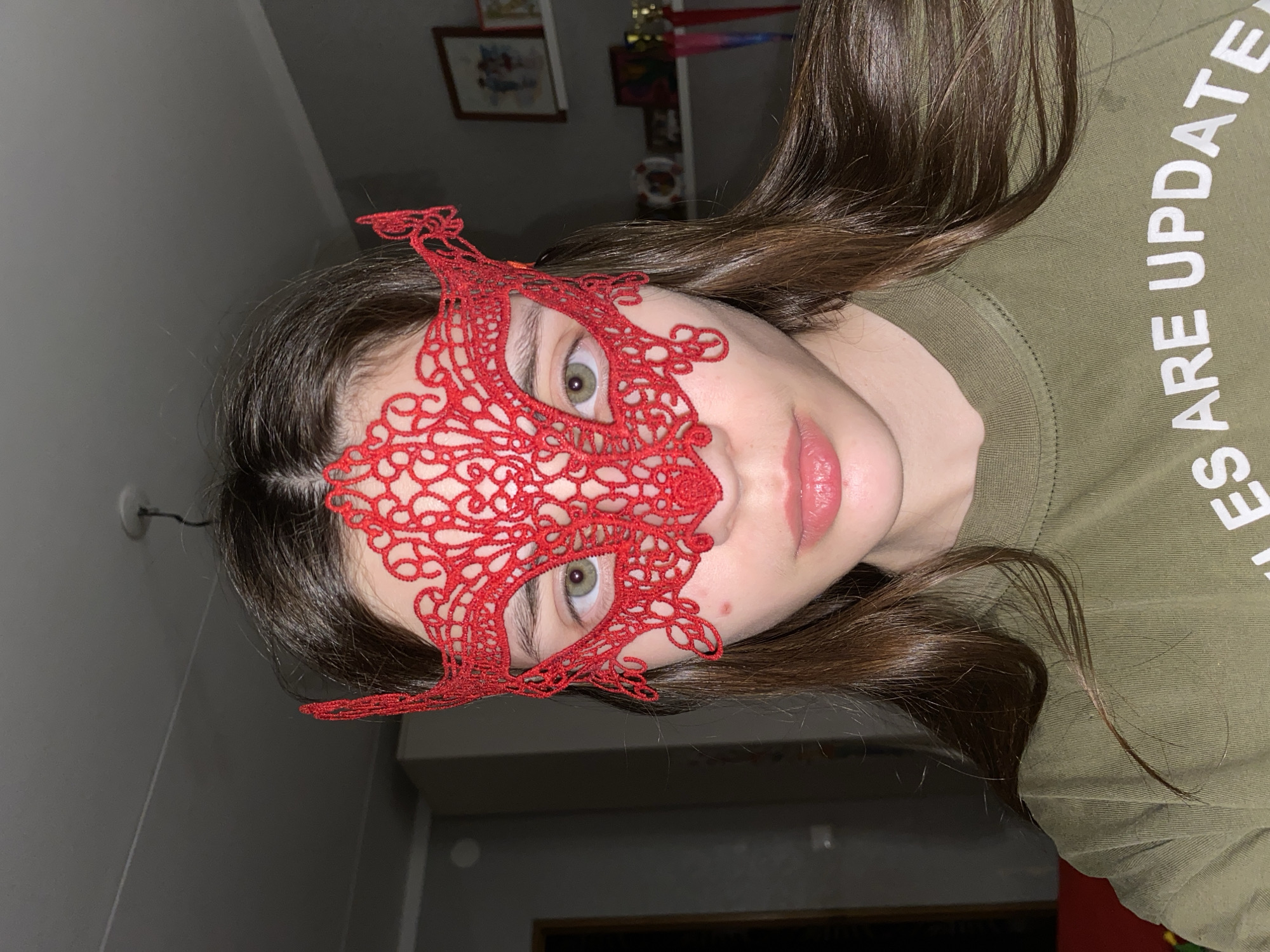 An indoor portrait-style photograph, taken in Votkinsk, Russia, features a young individual, likely female, with long, straight dark brown hair. The person's head is tilted to the side, and they are looking directly at the camera with green/hazel eyes. A prominent feature is a bright red, intricately patterned lace mask covering the upper half of their face, partially obscuring the forehead and nose, but leaving the eyes exposed. Their lips are a soft pink color, and a small red blemish is visible on the right cheekbone.
The individual is wearing an olive green long-sleeved shirt. White text is visible on the shoulder area of the shirt, reading "...S ARE UPDATED". The background is dimly lit, with dark gray walls. Visible background elements include framed pictures hanging on the wall, one depicting a cartoon-like image, and some shiny, colorful objects that resemble trophies or awards. The overall lighting suggests an indoor setting, possibly in the evening or with artificial illumination focused on the subject.