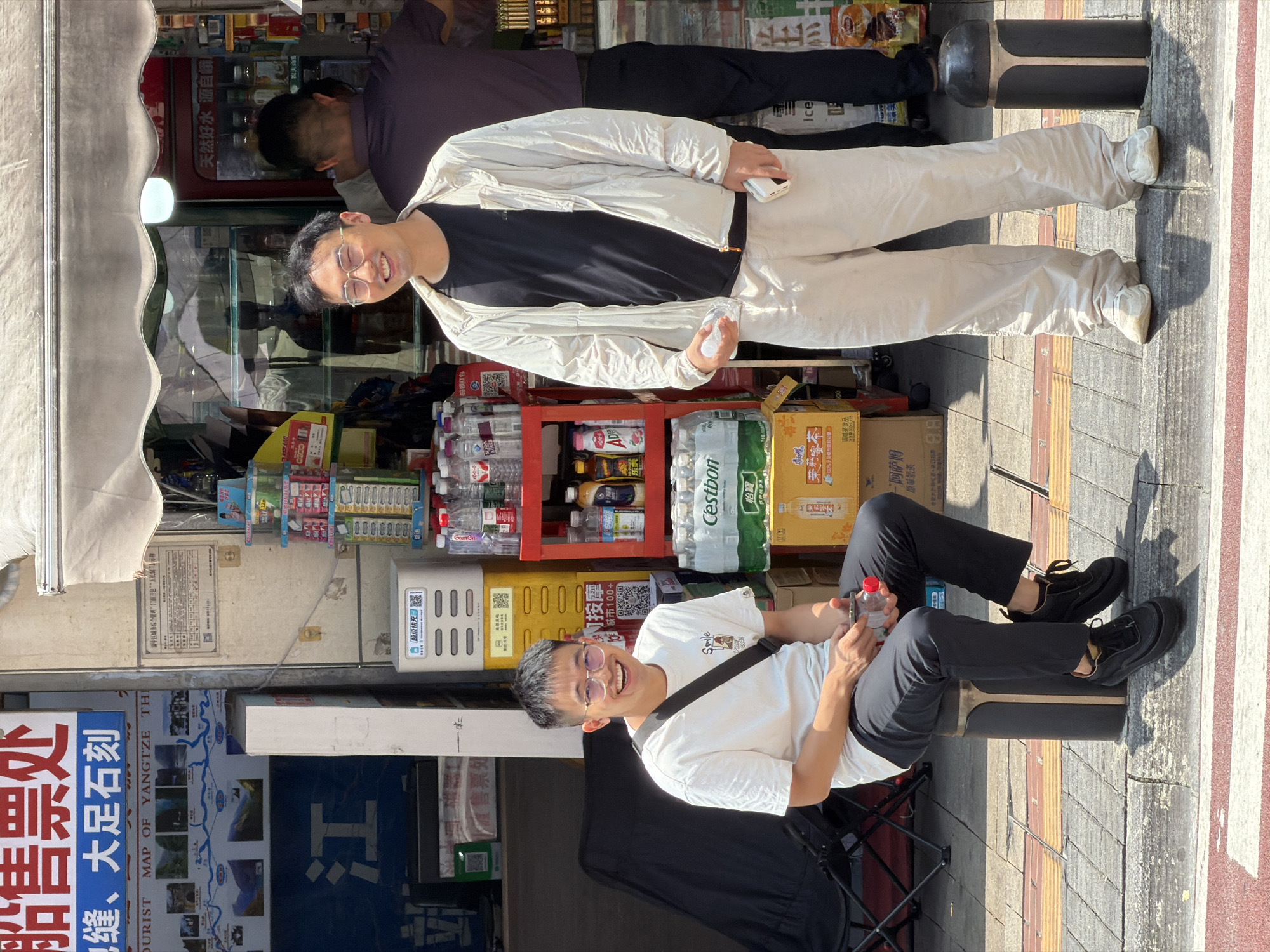 The image depicts an outdoor scene in front of a shop in Yuzhong District, China, on a sunny day. Two men are the primary subjects. The man on the left is seated on a dark cylindrical stool, wearing a white t-shirt, black pants, and black sneakers. He has short dark hair and glasses and is smiling. He holds a clear plastic bottle in his right hand. A black crossbody bag is slung over his shoulder.
The man on the right is standing, wearing a light-colored zip-up jacket over a black t-shirt, light-colored pants, and white sneakers. He also has short dark hair and glasses and is smiling. He holds a clear plastic bottle in his left hand and a white object, possibly a phone, in his right hand.
Behind the men, the shop entrance is visible. There are shelves stocked with various products, including bottled drinks and snacks. A poster with Chinese text and a map is displayed on the wall to the left. Another person, wearing a dark purple shirt, is partially visible in the background, facing away from the camera. The environment appears to be a street with a sidewalk, indicated by the paving stones and a curb.