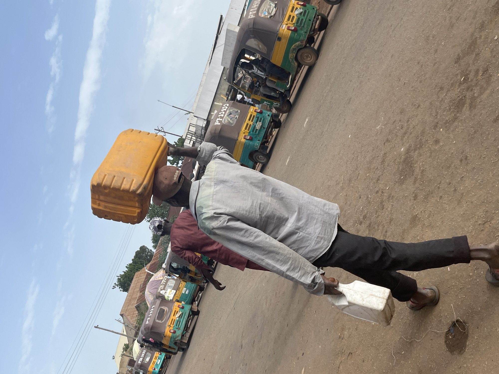 In Gura Top, Nigeria, a man in a grey collared shirt and dark pants carries an orange plastic container on his head and a white plastic jerrycan in his right hand. He is walking on a dirt and asphalt road. Behind him, another person in a maroon robe walks away from the camera. Several multi-colored auto-rickshaws are parked or driving on the road in the background. The sky is blue with scattered clouds. The scene appears to be a busy marketplace or transportation hub.