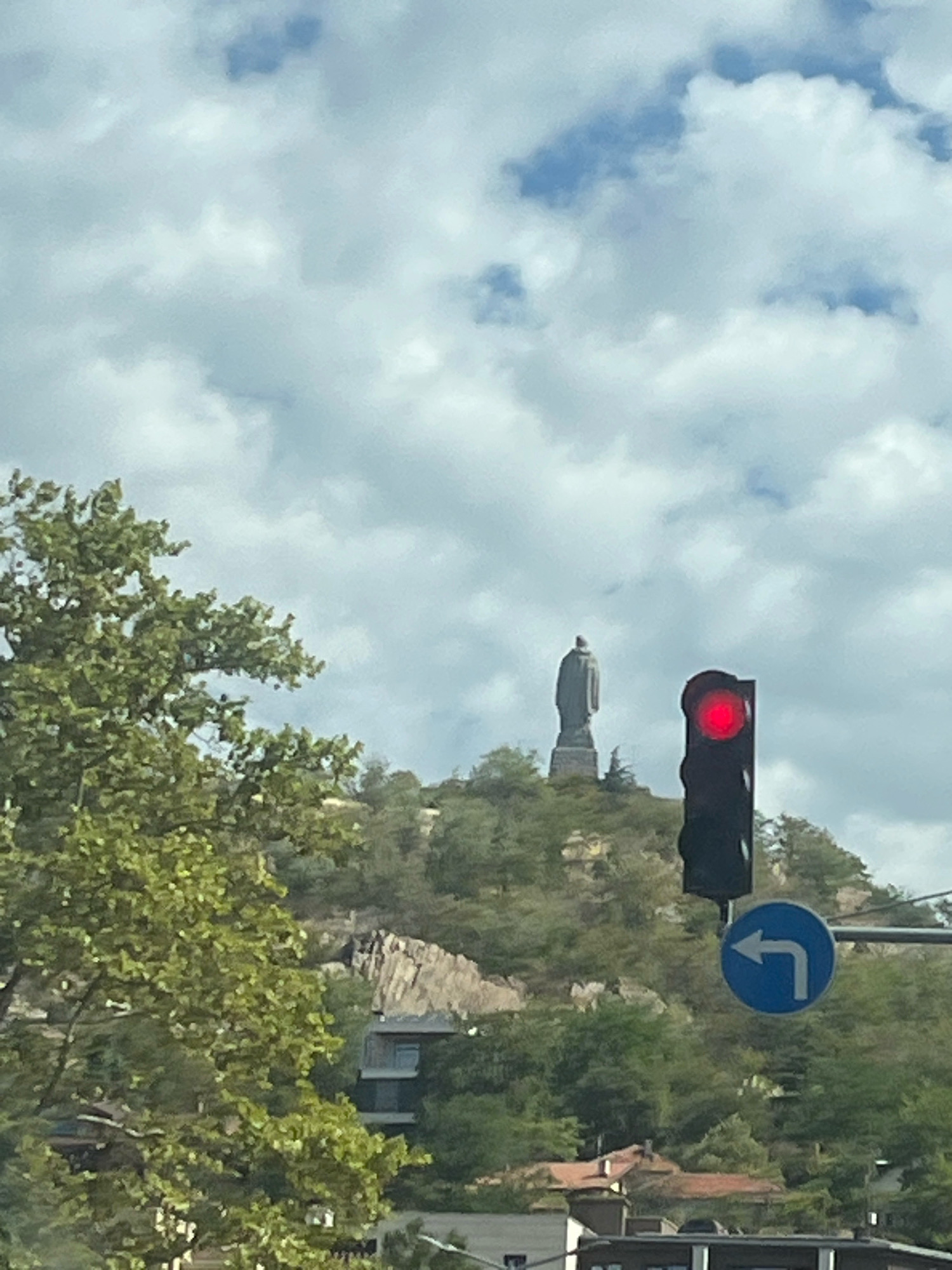 The image captures a view from a street level, likely within the city of Plovdiv, Bulgaria, as indicated by the presence of the iconic Alyosha Monument atop Bunardzhik Hill. In the foreground, a traffic light displays a red signal, and a blue circular sign with a white left-pointing arrow is visible below it, indicating a mandatory left turn. Lush green trees dominate the left side of the frame, obscuring some of the background. Further into the scene, Bunardzhik Hill rises, crowned by the imposing Alyosha Monument, a Soviet-era statue dedicated to the soldiers who fought in World War II. The hill is covered in dense vegetation and rocky outcrops. Several buildings, including some with red-tiled roofs, are nestled at the base of the hill and further in the background, suggesting an urban setting. The sky is partly cloudy, with white and grey clouds scattered against a blue backdrop. The overall impression is a typical street view with a significant landmark in the distance.