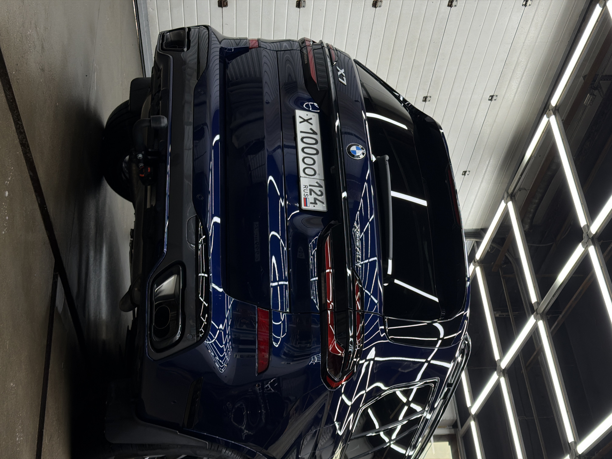 A dark blue BMW X7 is in a garage. The car's rear is facing the viewer. The license plate reads "X1000OO 124 RUS". The location is Krasnoyarsk, Russia. The garage has a white ceiling and bright fluorescent lights. The floor is gray concrete.