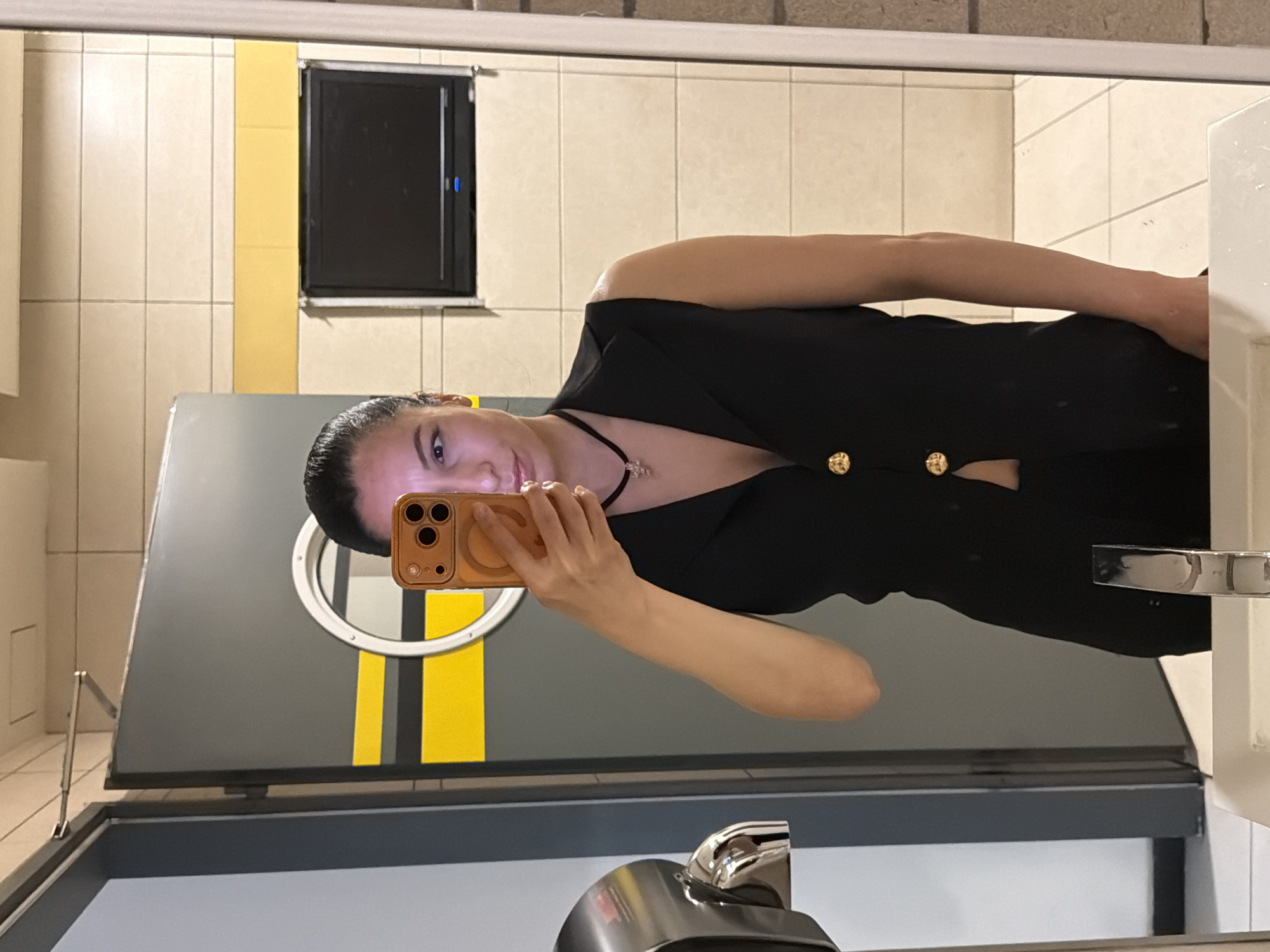 A woman is taking a selfie in a bathroom mirror. She is wearing a black top with gold buttons and a necklace. The bathroom has beige tile walls, a black wall-mounted appliance, a grey vanity with a round mirror and yellow stripe, and a stainless steel soap dispenser. The location appears to be a public restroom. There is no visible information to connect the image to the city of Pleven, Bulgaria.