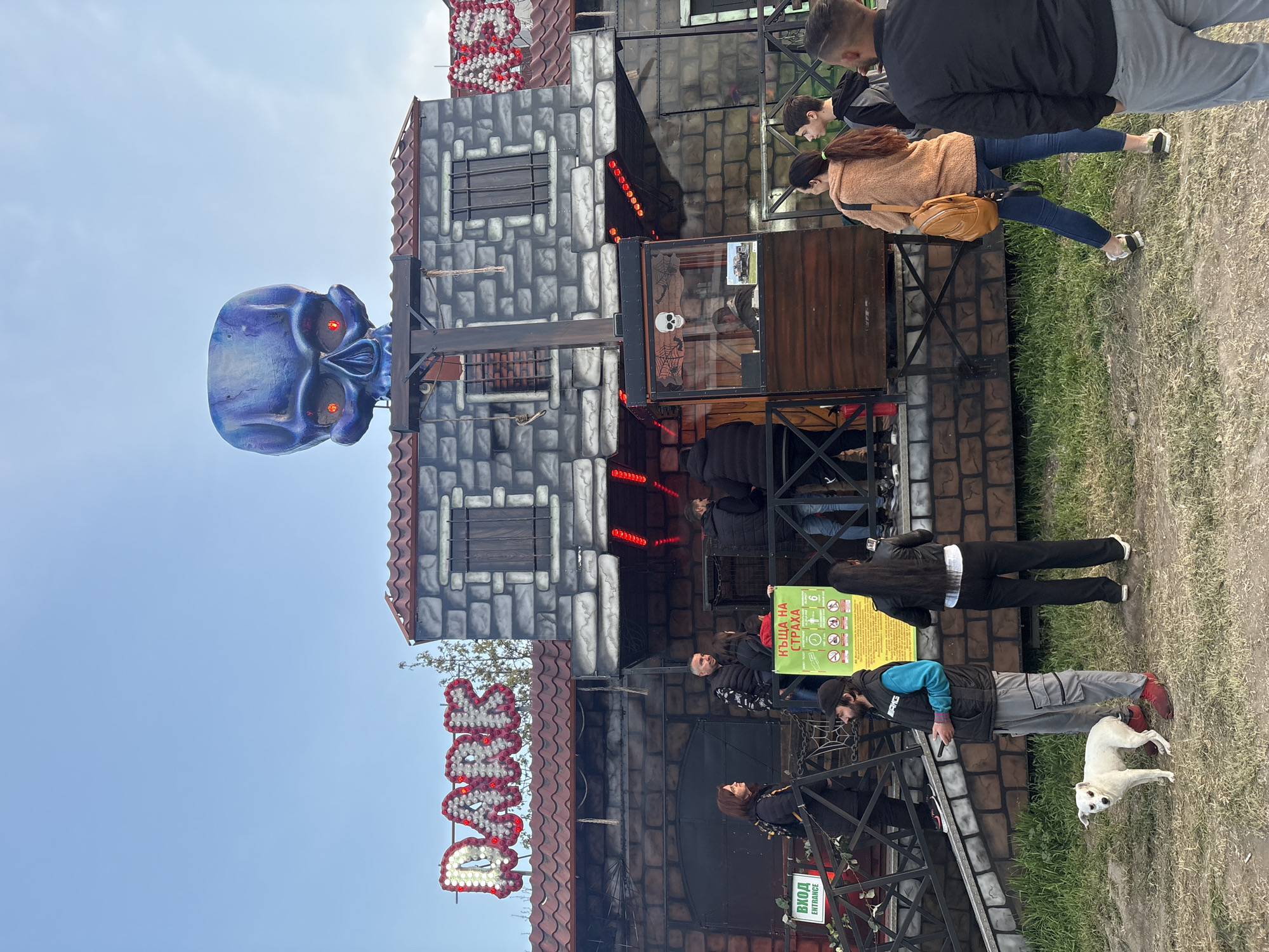 The image depicts a "DARK" house attraction in an unknown city. The structure is designed to resemble a stone building with a large blue skull with glowing red eyes mounted on top. Several people are standing around the entrance, including a man with a dog. There is a sign in a foreign language. The environment appears to be an outdoor fair or amusement park.