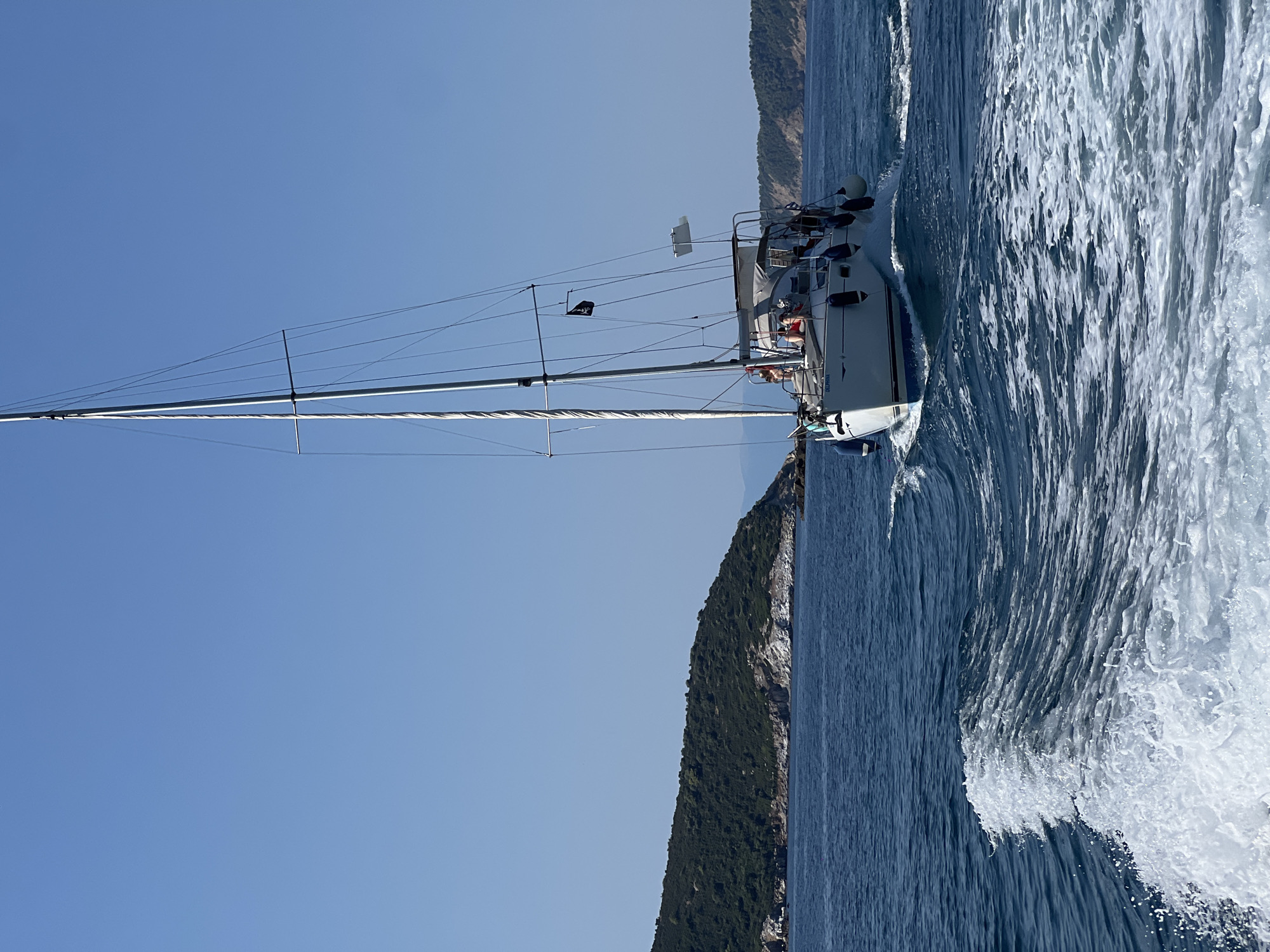 A white sailboat with a black skull and crossbones flag is sailing on the blue sea under a clear sky. The sailboat has several people on board. In the background, there is a green, rocky island with trees and a few buildings. The foreground shows white wake from the sailboat. The image appears to be taken from another boat moving in the same direction. The scene is likely located in the waters surrounding Skiathos, Greece, given the characteristic landscape of the Aegean islands.