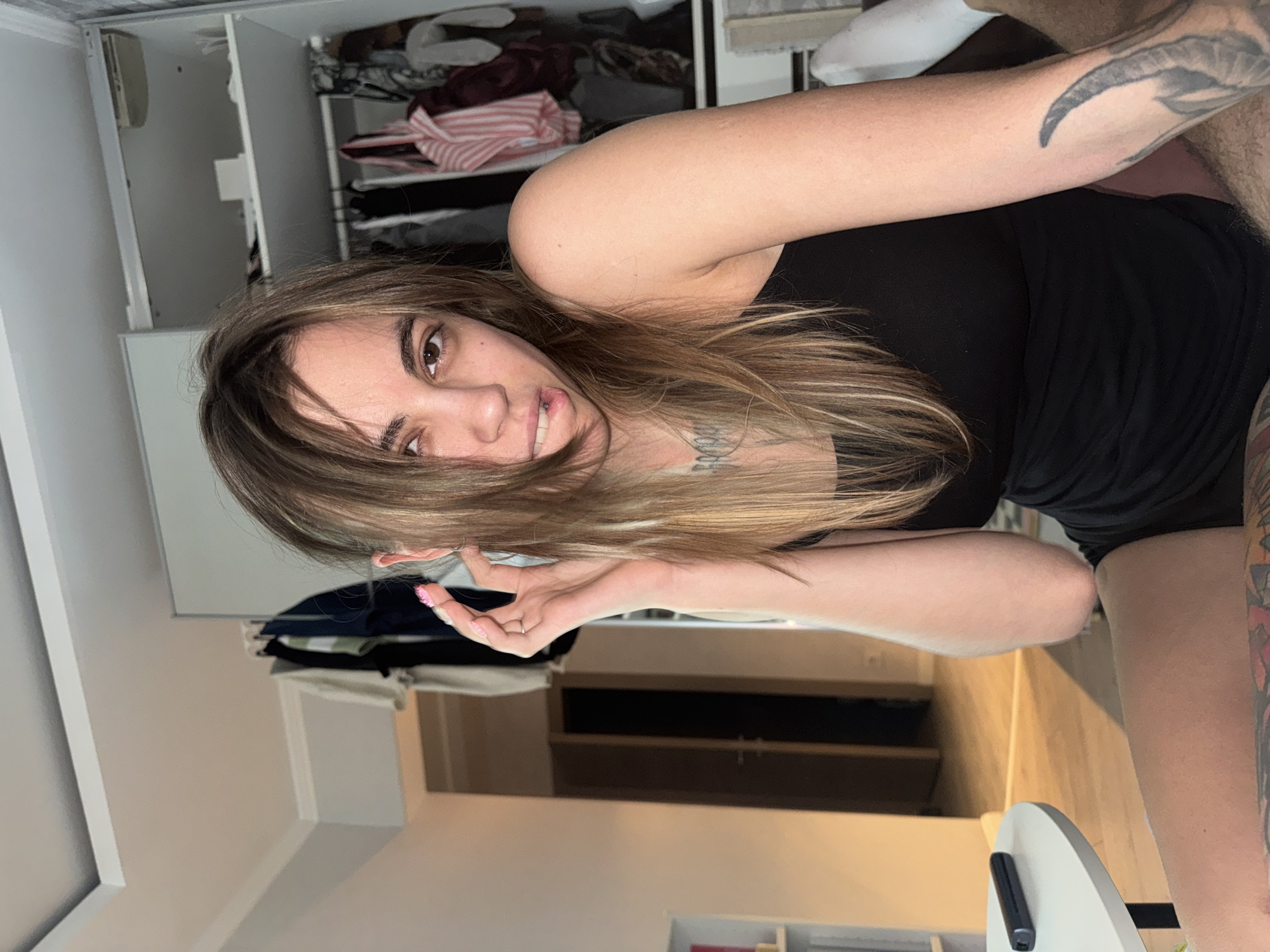 A young Caucasian woman with long, layered brown hair and light brown eyes is sitting in what appears to be a bedroom or dressing area. She is wearing a black tank top and black shorts. Her right hand is raised to her ear, and she is biting her lower lip. She has a lip piercing and tattoos on her neck and arm. In the background, there is an open wardrobe with clothes hanging inside, and a door in the hallway. On a round white table to her left, there is a black object, possibly a vape pen. The lighting suggests it is daytime, and the scene is indoors. There is no indication of the specific location within Moscow, Russia.