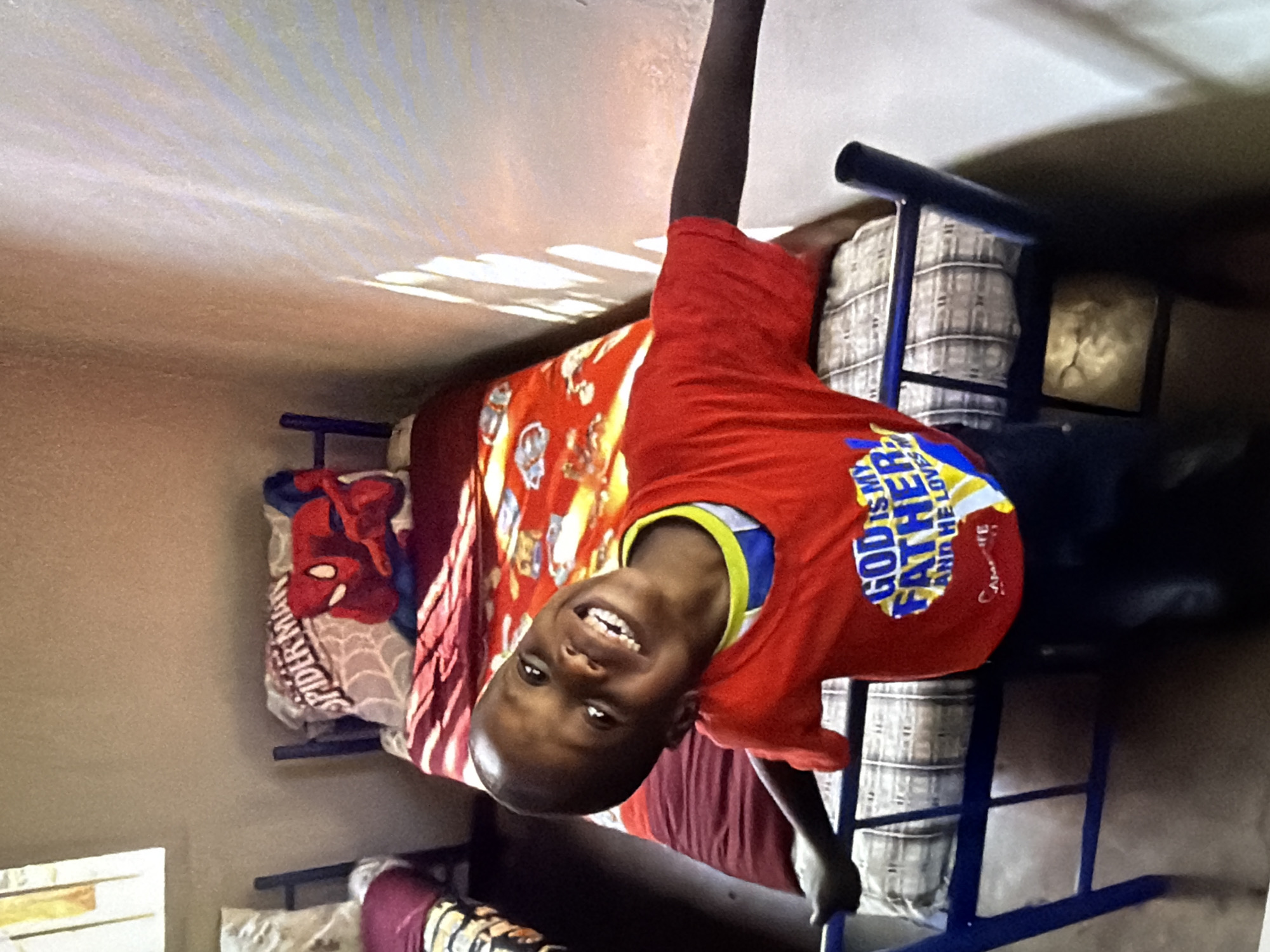 A young boy with dark skin and short hair is looking at the camera with a wide smile. He is wearing a red t-shirt with a yellow and blue collar that has the words "GOD IS MY FATHER AND HE LOVES ME" printed on it in yellow and blue letters. The boy is standing next to a bunk bed with a blue metal frame. The bed is covered with a red and white striped blanket and a pillow with a Spiderman design. The background of the image is a plain wall with sunlight streaming through a window, casting shadows on the wall. The scene appears to be indoors, possibly in a dormitory or shelter.
