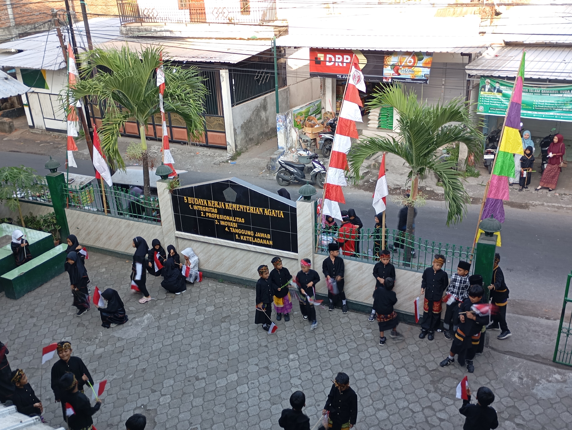 The image depicts an outdoor scene in Mataram, Indonesia, possibly in front of an educational institution, as indicated by the text on a plaque reading "5 BUDAYA KERJA KEMENTERIAN AGAMA" (5 Work Cultures of the Ministry of Religion). A group of children, some dressed in traditional Islamic attire including headscarves and sarongs, and others in black uniforms with red and white sashes or bands, are gathered on a paved area. Many of the children are holding small Indonesian flags, a symbol of national pride.
Several Indonesian flags are prominently displayed throughout the scene, some on tall poles and others handheld. In the background, there are buildings with metal gates, corrugated roofs, and shop fronts. Palm trees are also visible, contributing to the tropical environment. Motorbikes are parked near the buildings, suggesting a typical urban setting. The overall atmosphere appears to be a patriotic or celebratory event involving children. The pavement is patterned with interlocking gray stones. The time of day appears to be late afternoon, with warm lighting casting shadows.