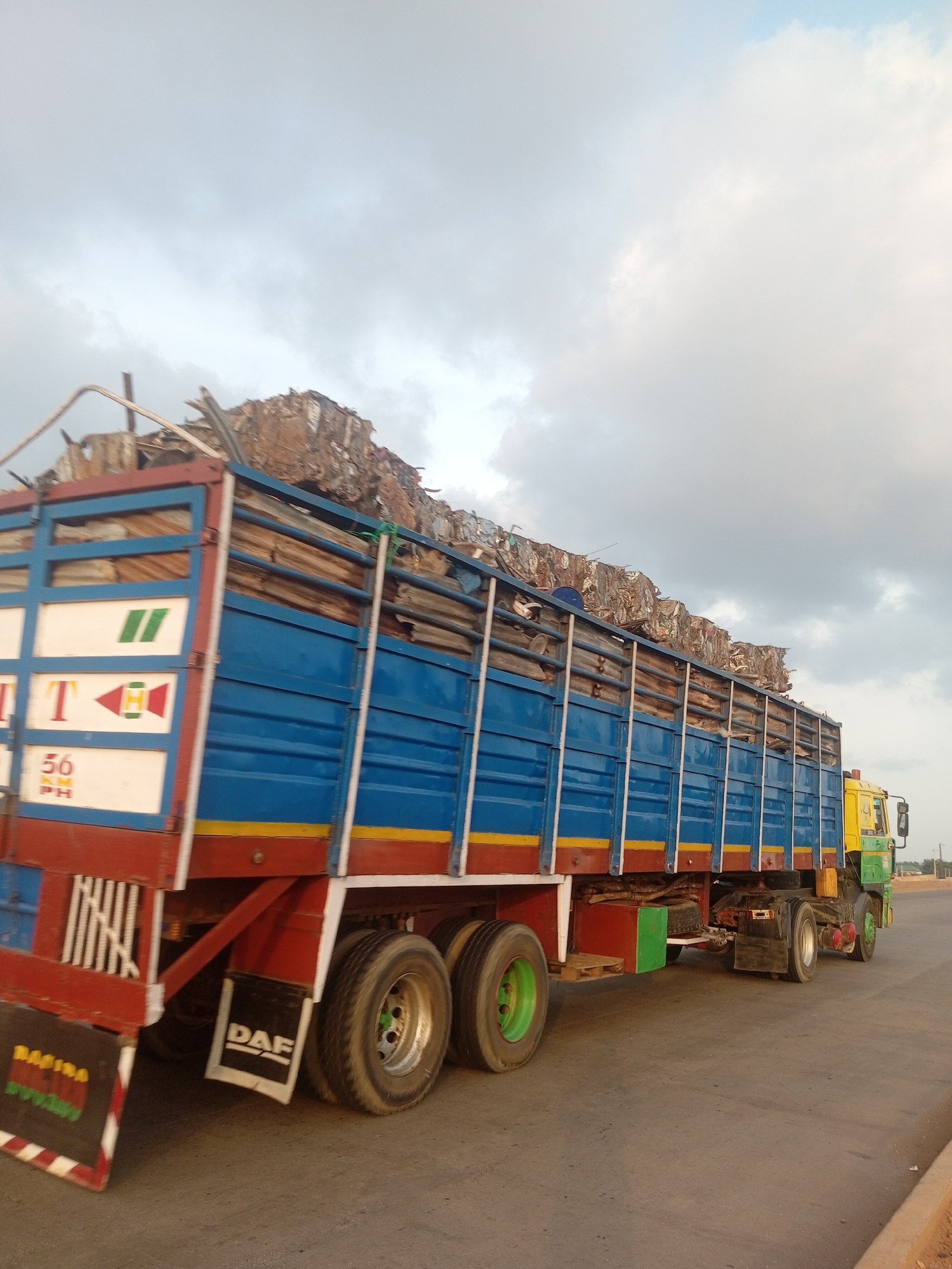 An eye-level, full shot captures a large, blue and red dump truck traveling on an asphalt road under a cloudy sky. The truck is loaded with a significant amount of scrap metal, which is piled high above the truck's bed. The truck has a yellow and green cab. The road appears to be in an undeveloped area with sparse vegetation visible in the background. The lighting suggests it is either early morning or late afternoon.