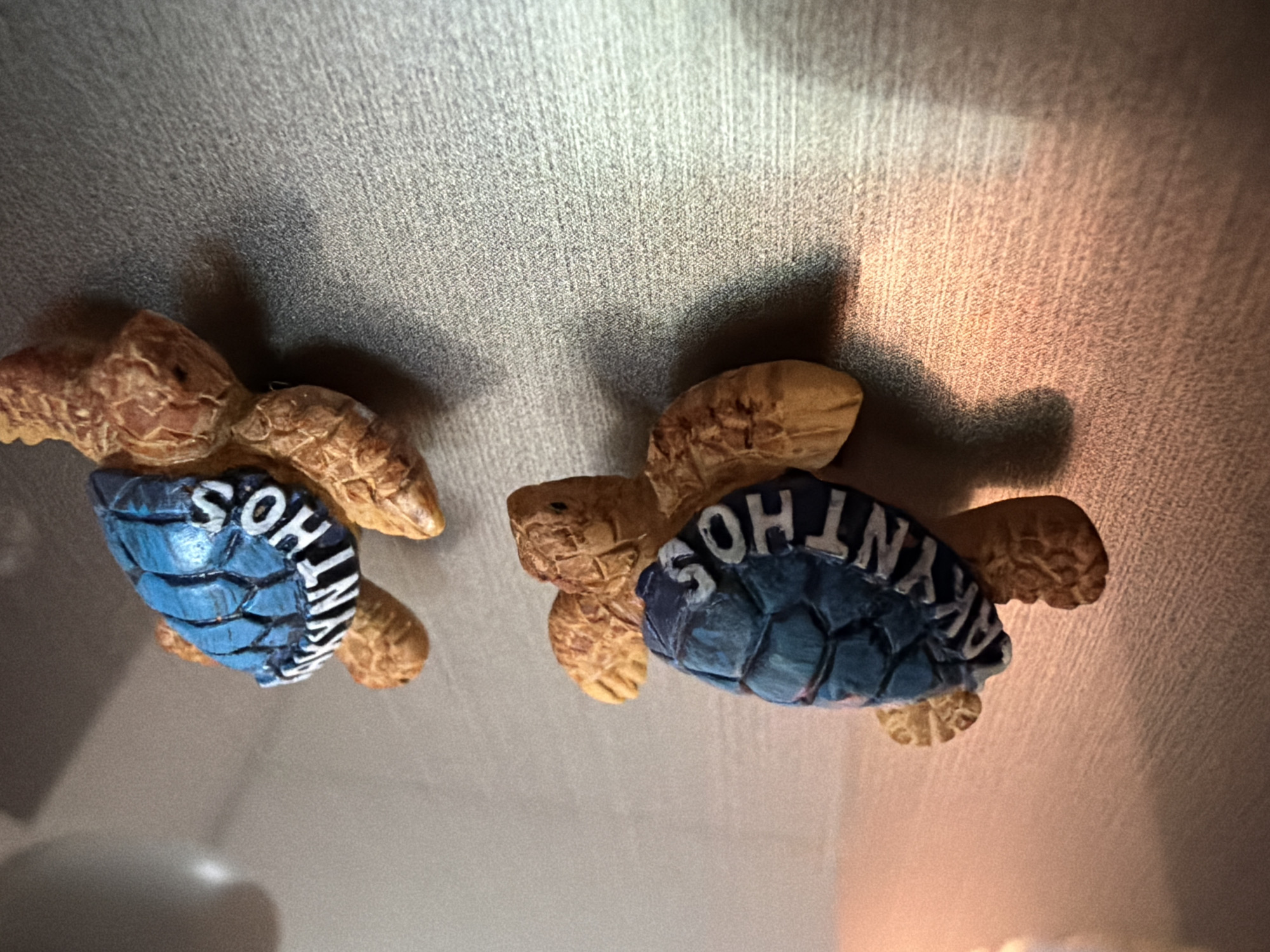Two decorative turtle figurines are displayed on a textured gray surface. The turtles are facing different directions, with their shells painted blue and embossed with white lettering that spells "MYKONOS". The top turtle is positioned higher in the frame and angled to the right, while the bottom turtle is lower and angled to the left. The surface they are on appears to be a magnetic board, indicated by the way the figurines are attached. A warm, soft light source is casting shadows from the turtles onto the surface to their right. The background is out of focus, with a blurred vertical line suggesting a wall or another surface. The overall scene is a close-up shot of these two souvenir items.