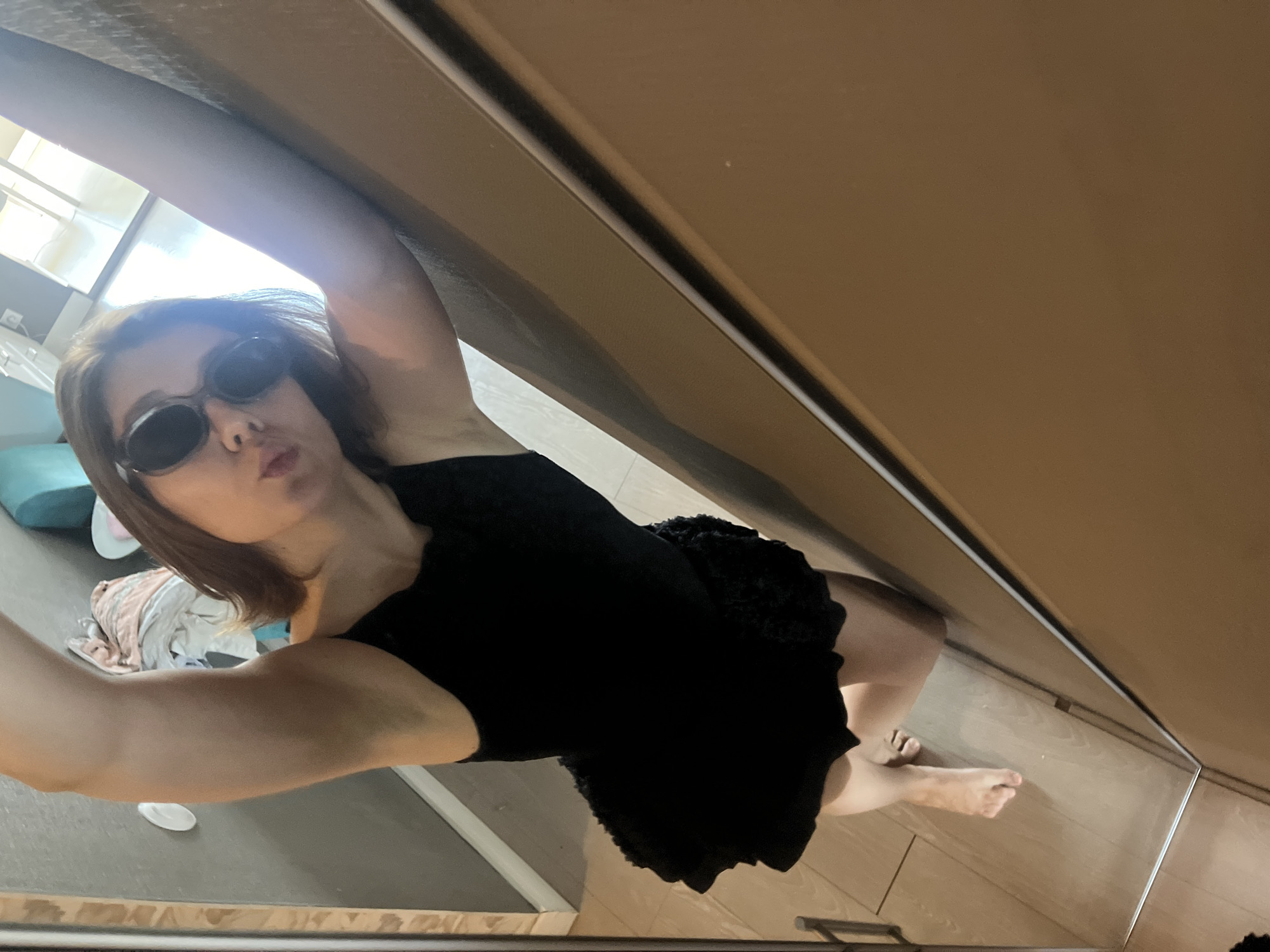 The image shows a young woman in a black dress and sunglasses taking a selfie in a mirrored surface. She is leaning against a light-colored wall, with her left arm extended upwards. The background appears to be an indoor setting, possibly a home or dressing room, with some laundry visible in the background. The floor is made of wooden planks. There is no discernible information in the image that would suggest a location in Krasnoyarsk, Russia.